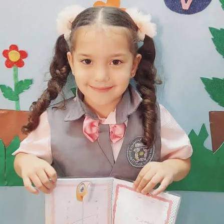 Hind Rajab in her school uniform — grey vest, pink shirt, pink bow tie — holding papers and smiling in her classroom.