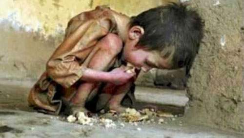 A malnourished child crouches against a wall, eating scraps of food off the ground.