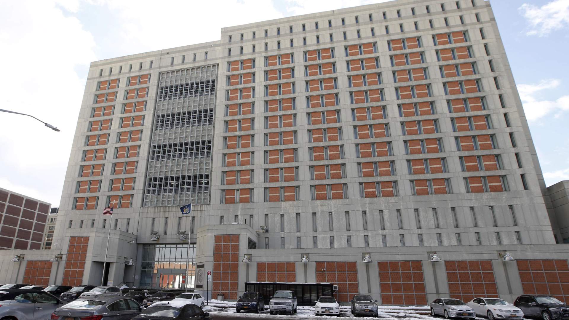 Exterior view of the Metropolitan Detention Center in Brooklyn, a large modern prison building with fencing along the street in Sunset Park