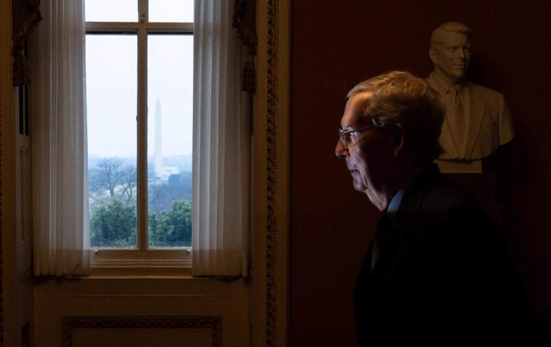 Mitch McConnell walking down a hallway carrying a binder, with American flags in the background