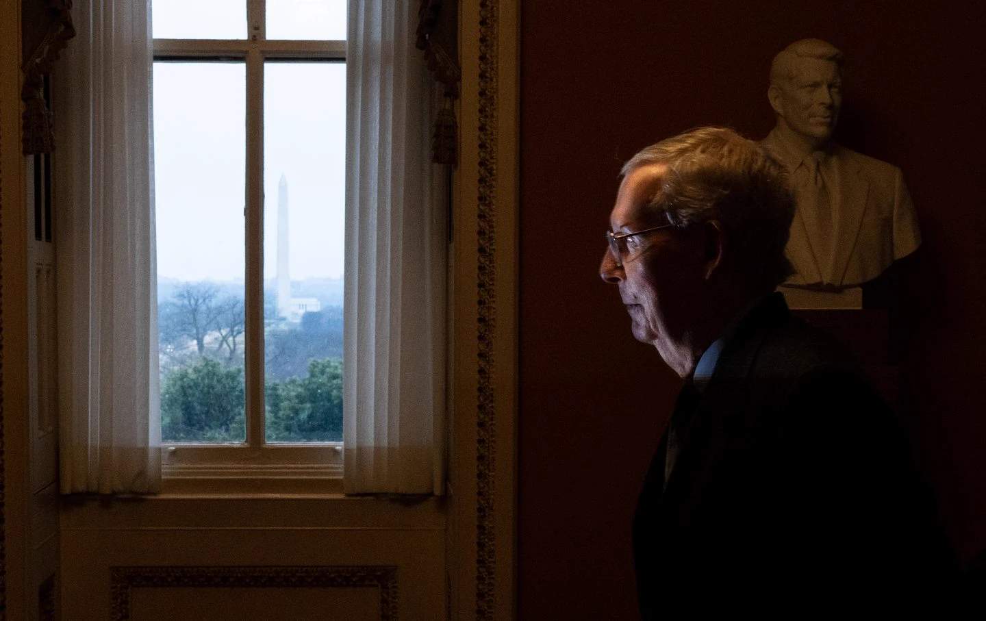 Mitch McConnell walking down a hallway carrying a binder, with American flags in the background