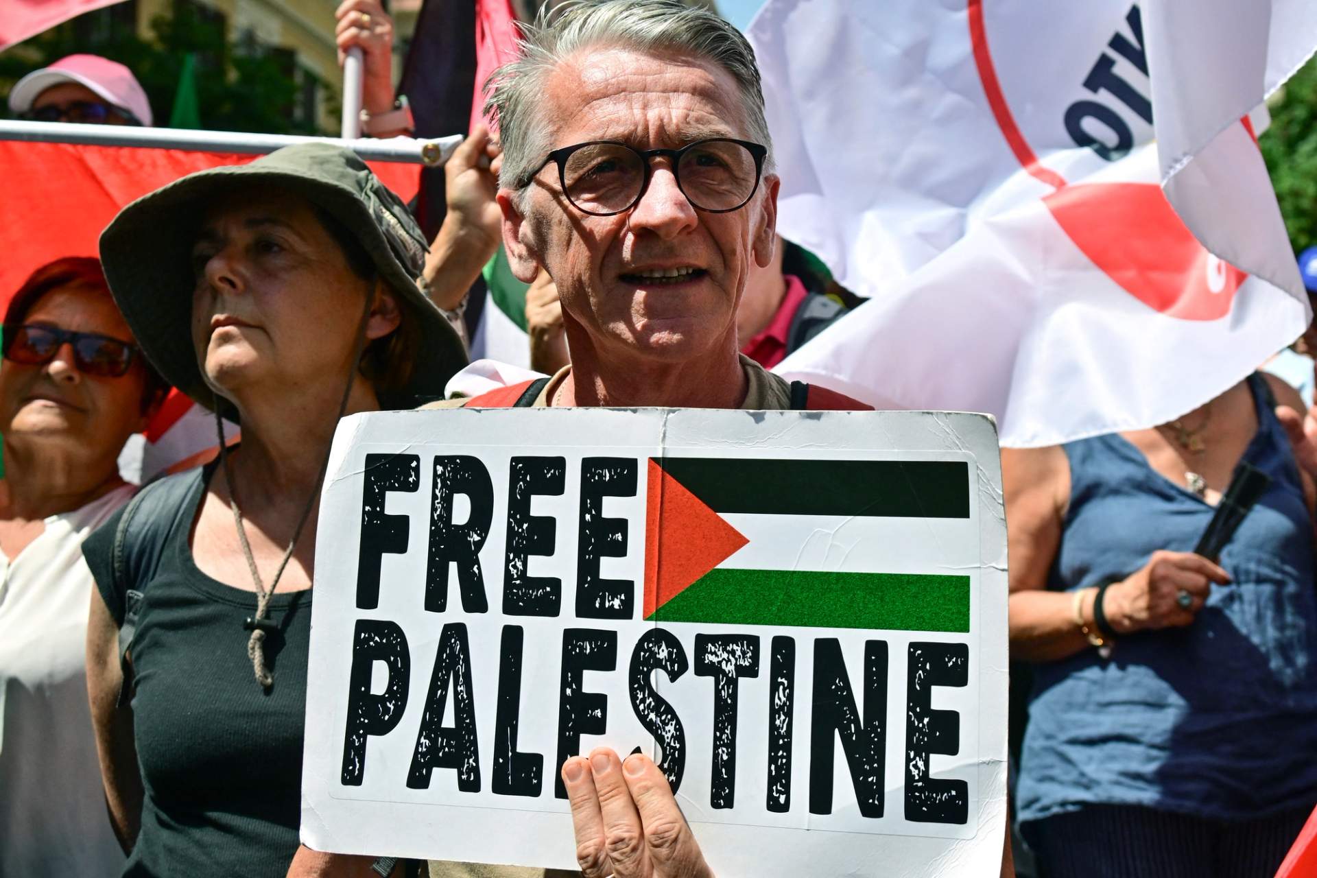 A crowd of protesters march through a city street, with one man prominently holding a white placard that reads 'Free Palestine' in black letters. Other protesters wave Palestinian flags and signs in the background, under a blue sky with scattered clouds.