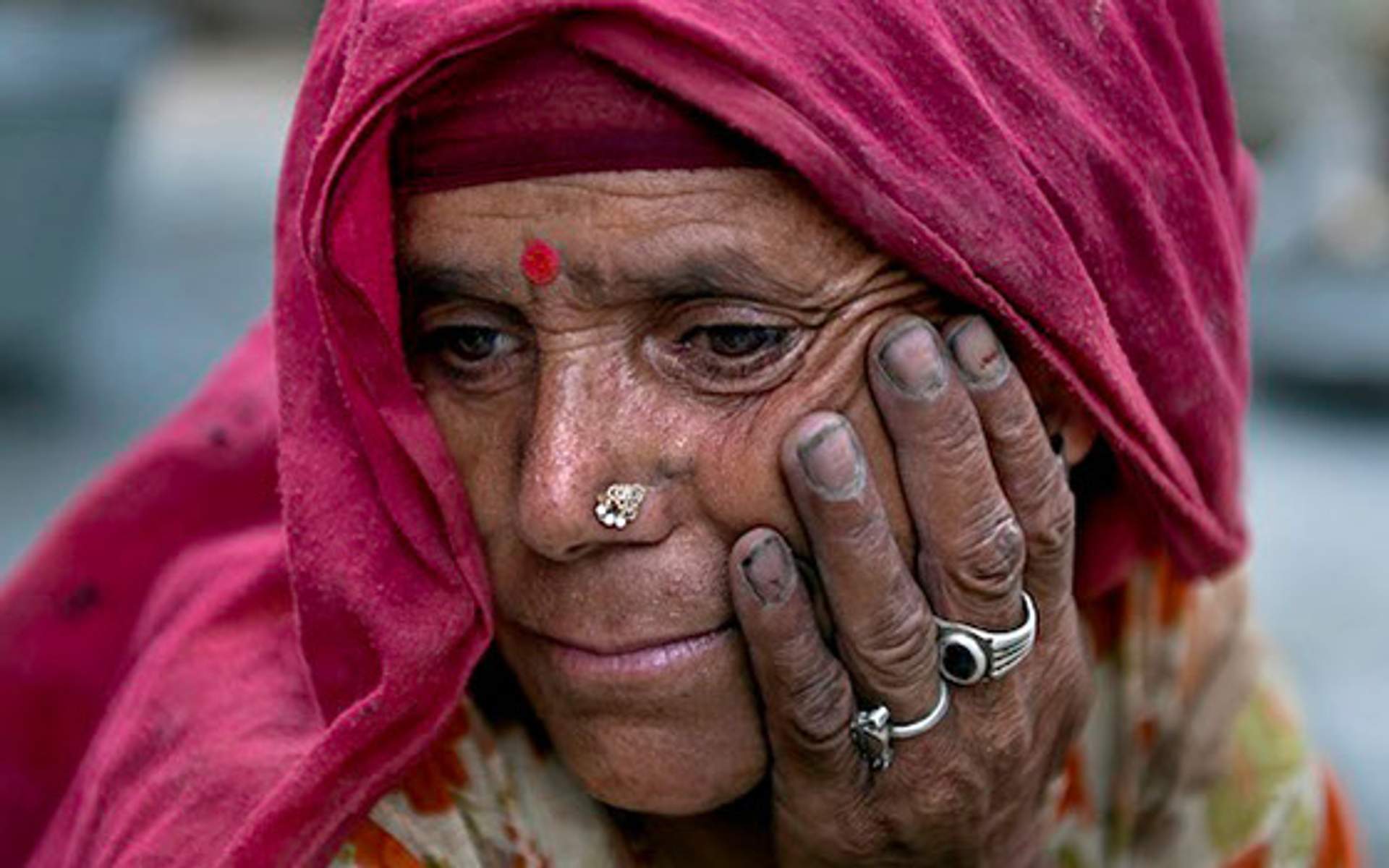 An elderly woman in a red headscarf rests her face in her hand.