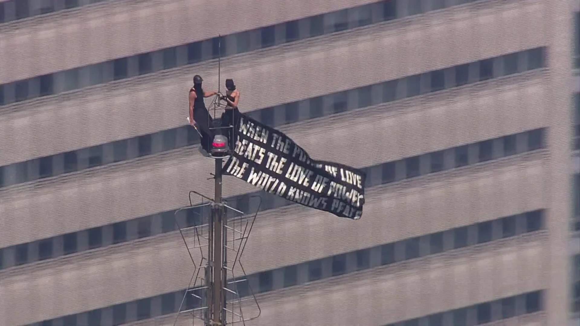 Two masked climbers perch on the Empire State Building antenna spire, holding a black-and-white peace banner, 1,454 feet above Midtown Manhattan on July 1, 2026.