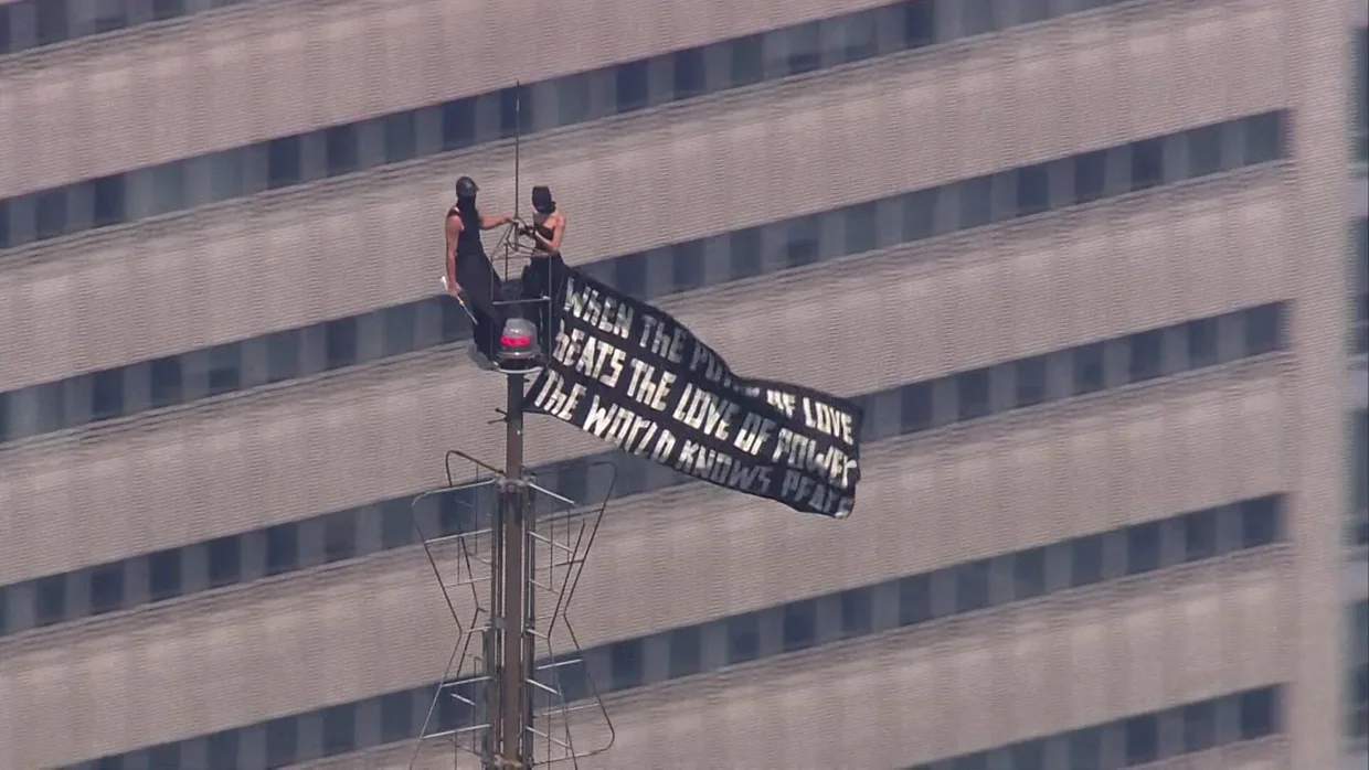 Two masked climbers perch on the Empire State Building antenna spire, holding a black-and-white peace banner, 1,454 feet above Midtown Manhattan on July 1, 2026.