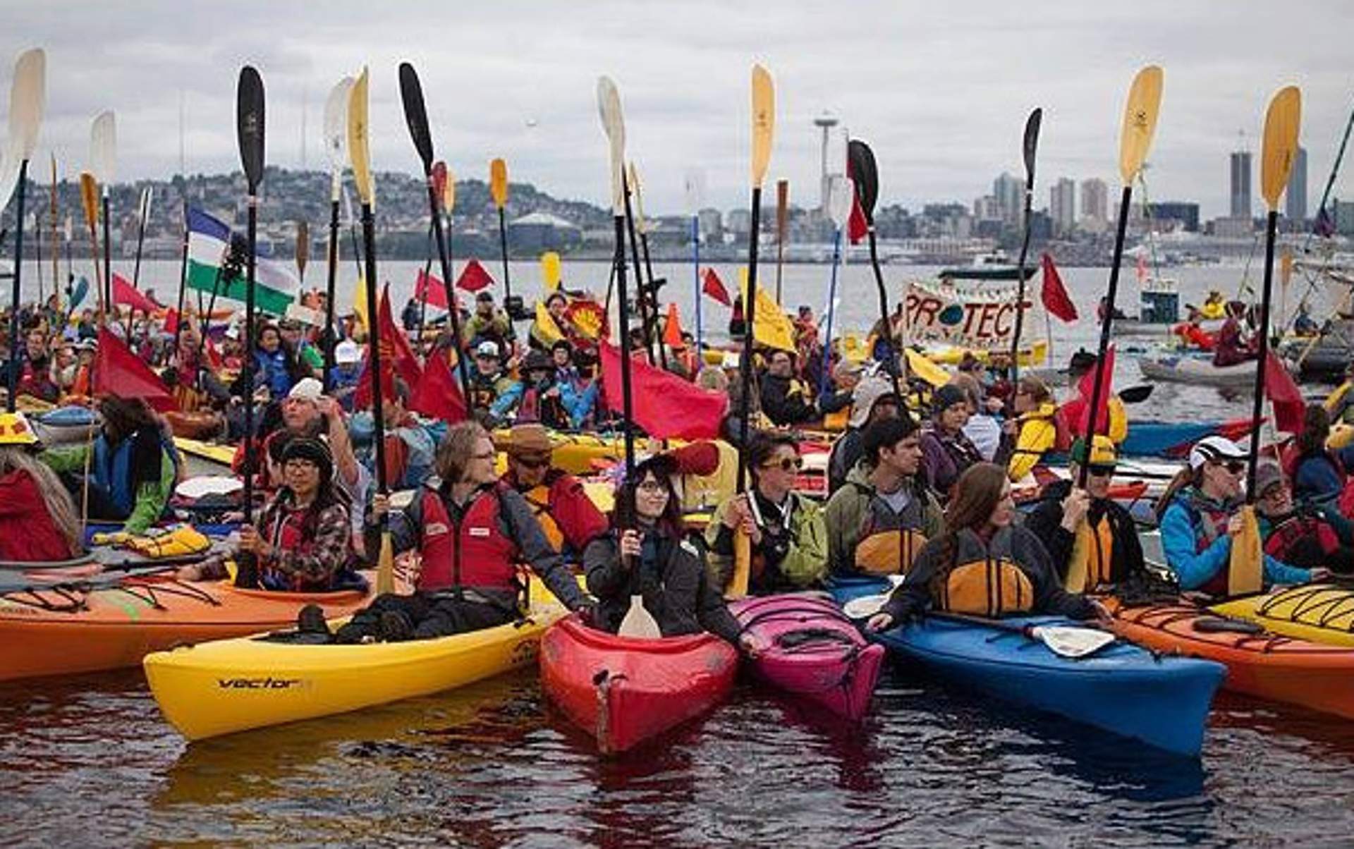 Dozens of kayakers holding paddles and flags gather on the water with the Seattle skyline in the background.