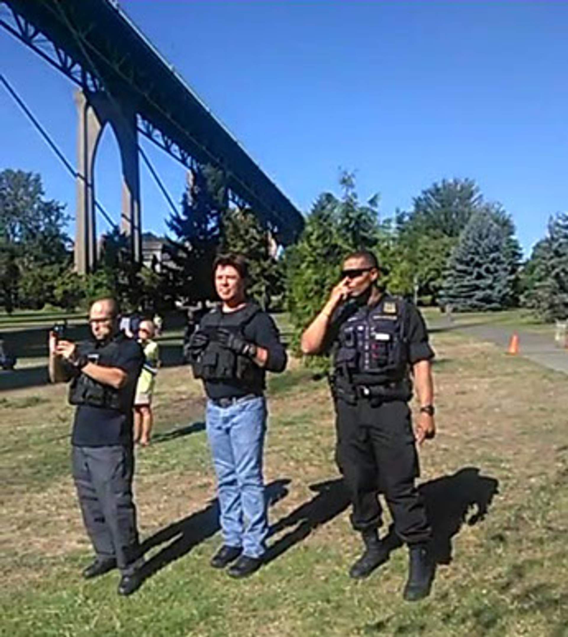 Three men in tactical vests stand near the bridge, one holding a camera and another speaking on a radio.