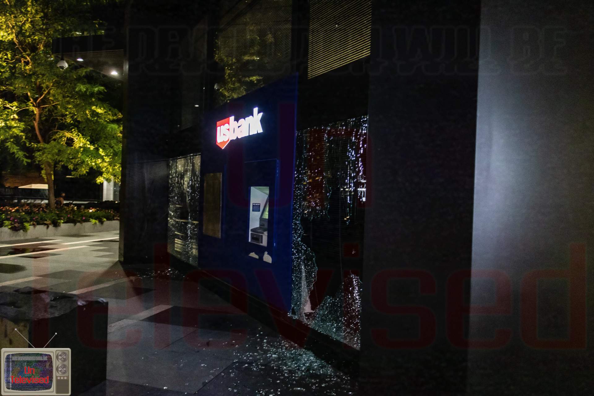 A U.S. Bank branch/ATM kiosk with its glass panel smashed, shards scattered on the sidewalk below the lit "US Bank" sign.  [Radical Edward - Untelevised Media]