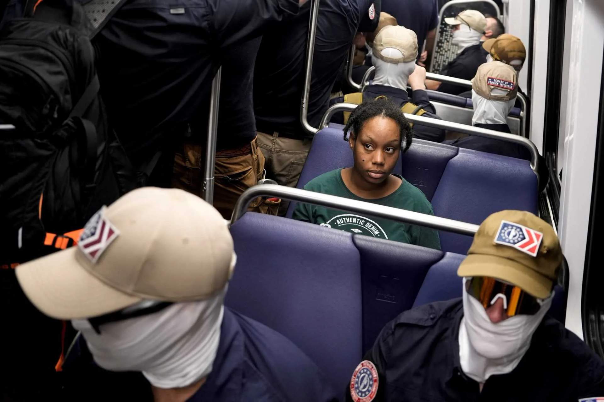 Multiple masked men in matching navy blue shirts fill a Metro train car, standing and holding onto overhead rails, while other passengers sit nearby