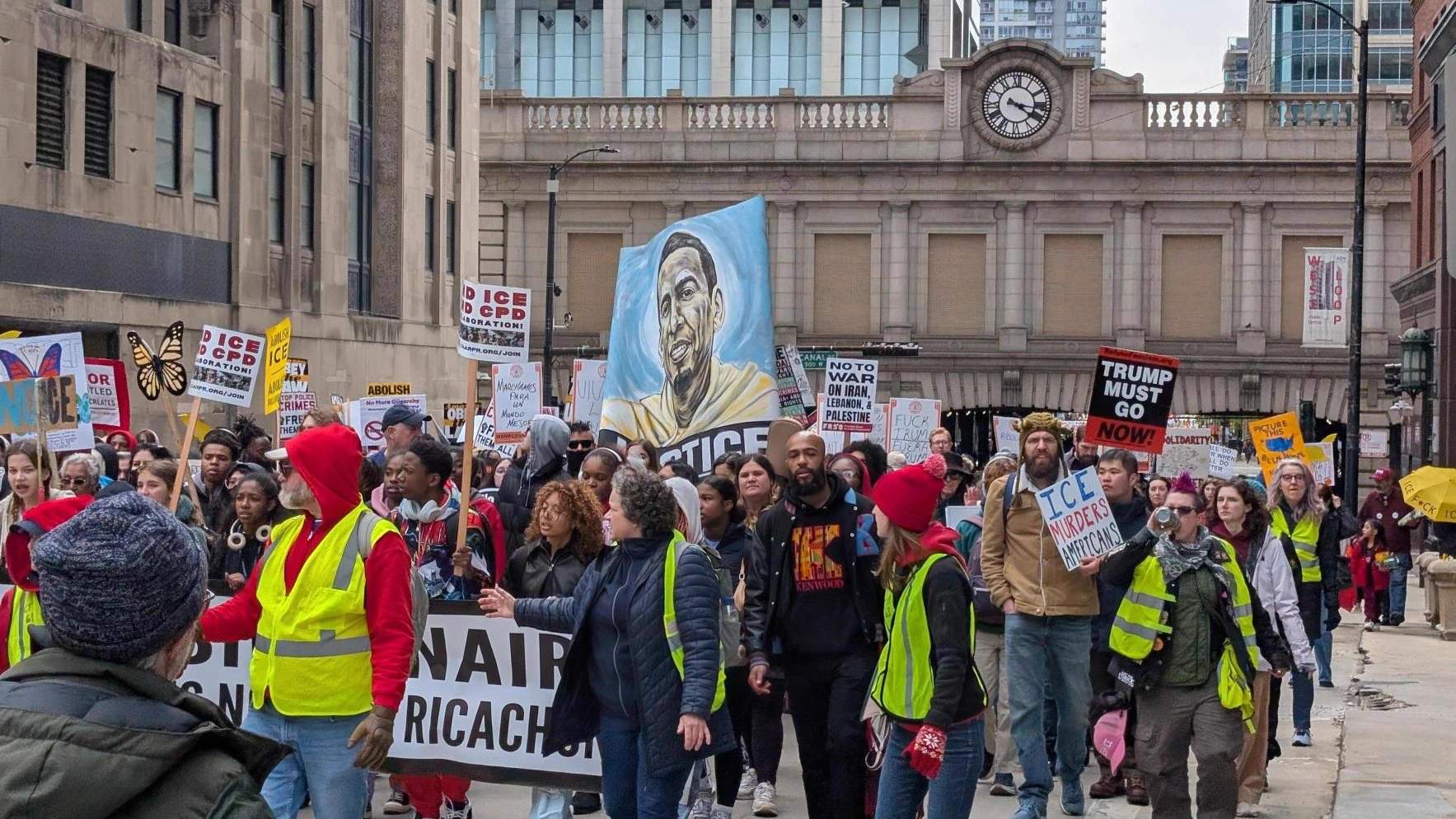 Large crowd of demonstrators marching down a city street carrying signs and banners during a May Day labor rally in Chicago, with buildings visible on both sides