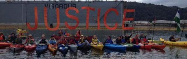 Kayaktivists in a row of boats hold up a giant orange 'JUSTICE' banner on the water beneath a bridge.