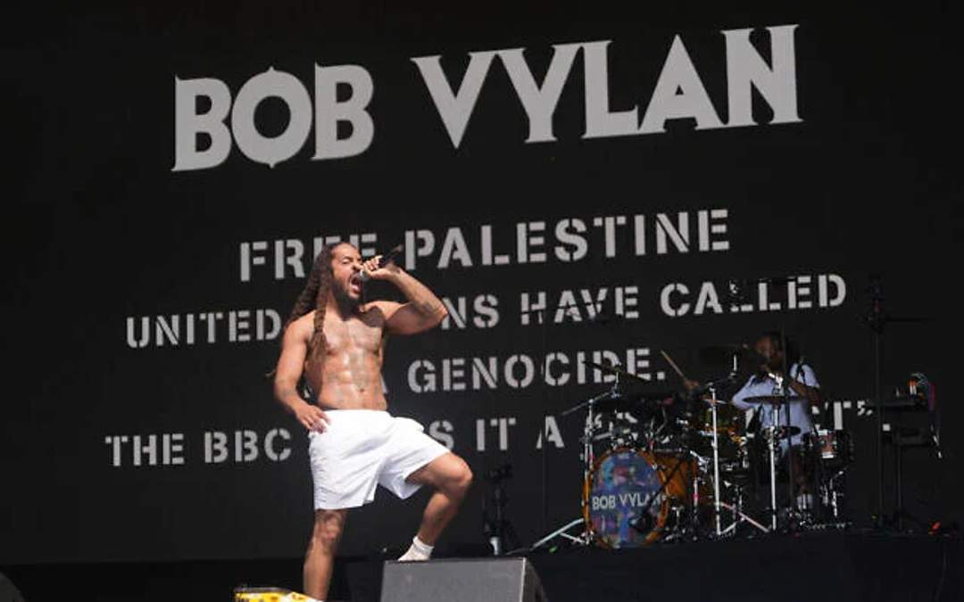 Bob Vylan, a Black man with dreadlocks, performs on a large outdoor stage at Glastonbury Festival, holding a microphone and engaging with the crowd. Behind him, festival-goers wave Palestinian flags and respond to his performance.