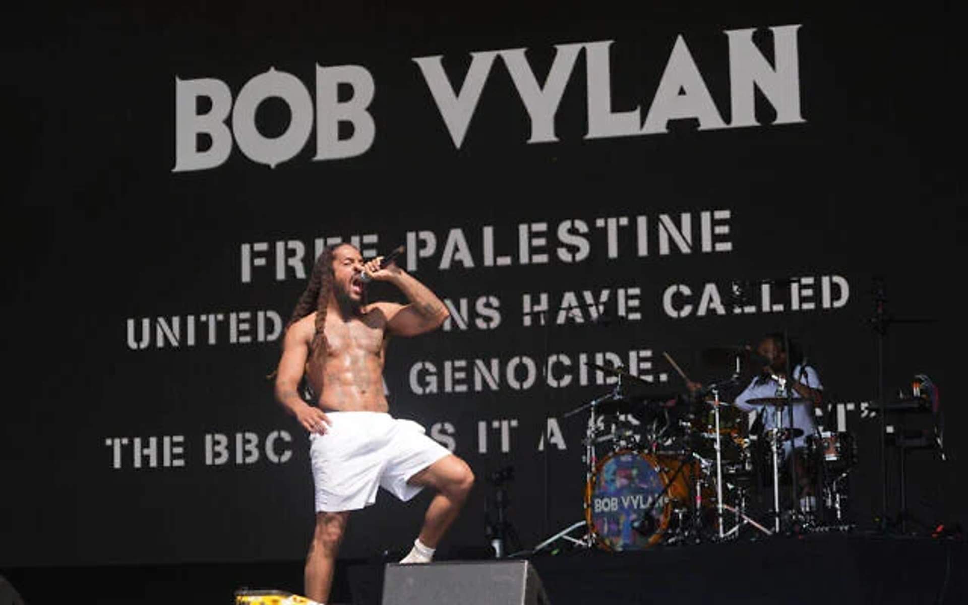 Bob Vylan, a Black man with dreadlocks, performs on a large outdoor stage at Glastonbury Festival, holding a microphone and engaging with the crowd. Behind him, festival-goers wave Palestinian flags and respond to his performance.