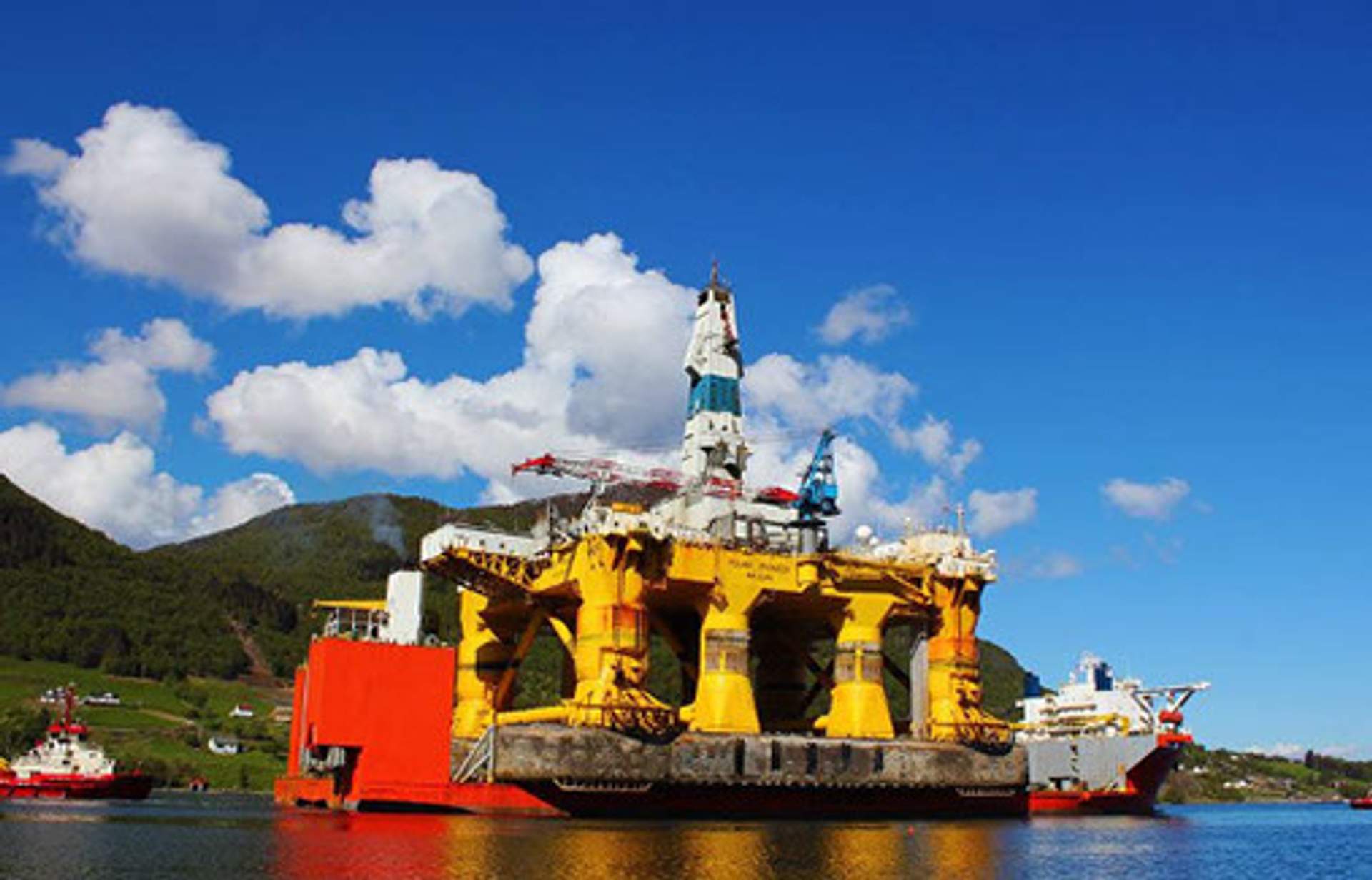 The Polar Pioneer, a massive yellow semi-submersible drilling rig, docked near green hills under a blue sky.