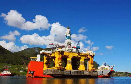 The Polar Pioneer, a massive yellow semi-submersible drilling rig, docked near green hills under a blue sky.