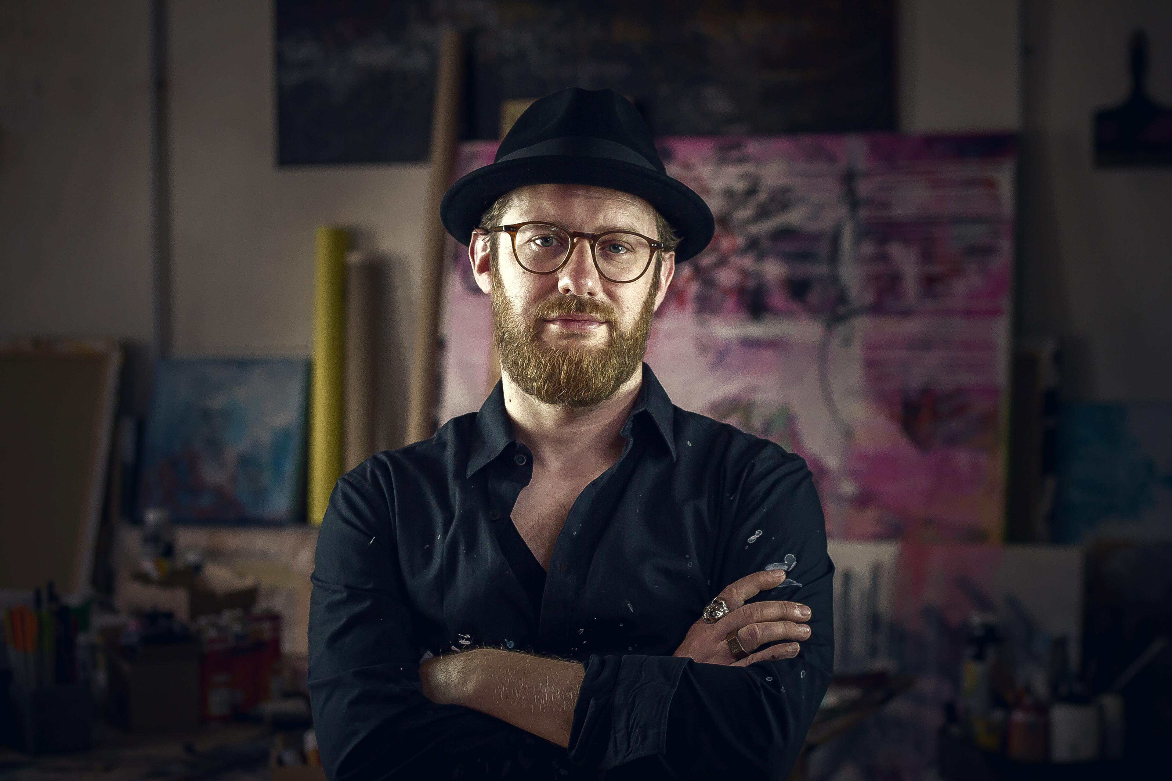 Portraitfoto des Künstler Patrick Hartl, mit verschränkten Armen in seinem Studio vor Arbeiten im Hintergrund