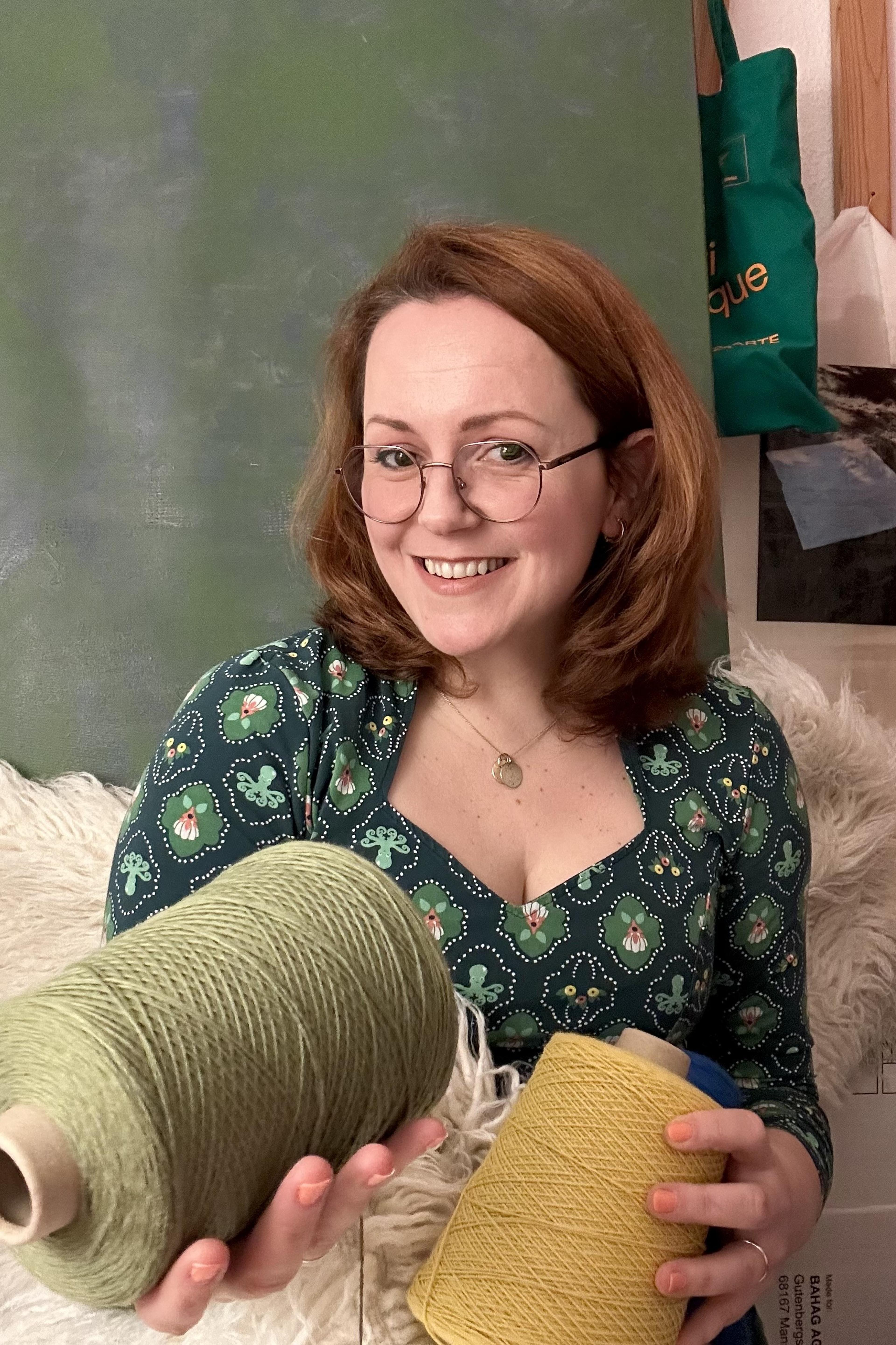 A woman with red, shoulder length hair sits on a furry chair. She wears a green blouse and holds two wool spouls in her hands. She looks at the camera and smiles. In the background there is a light green wall.