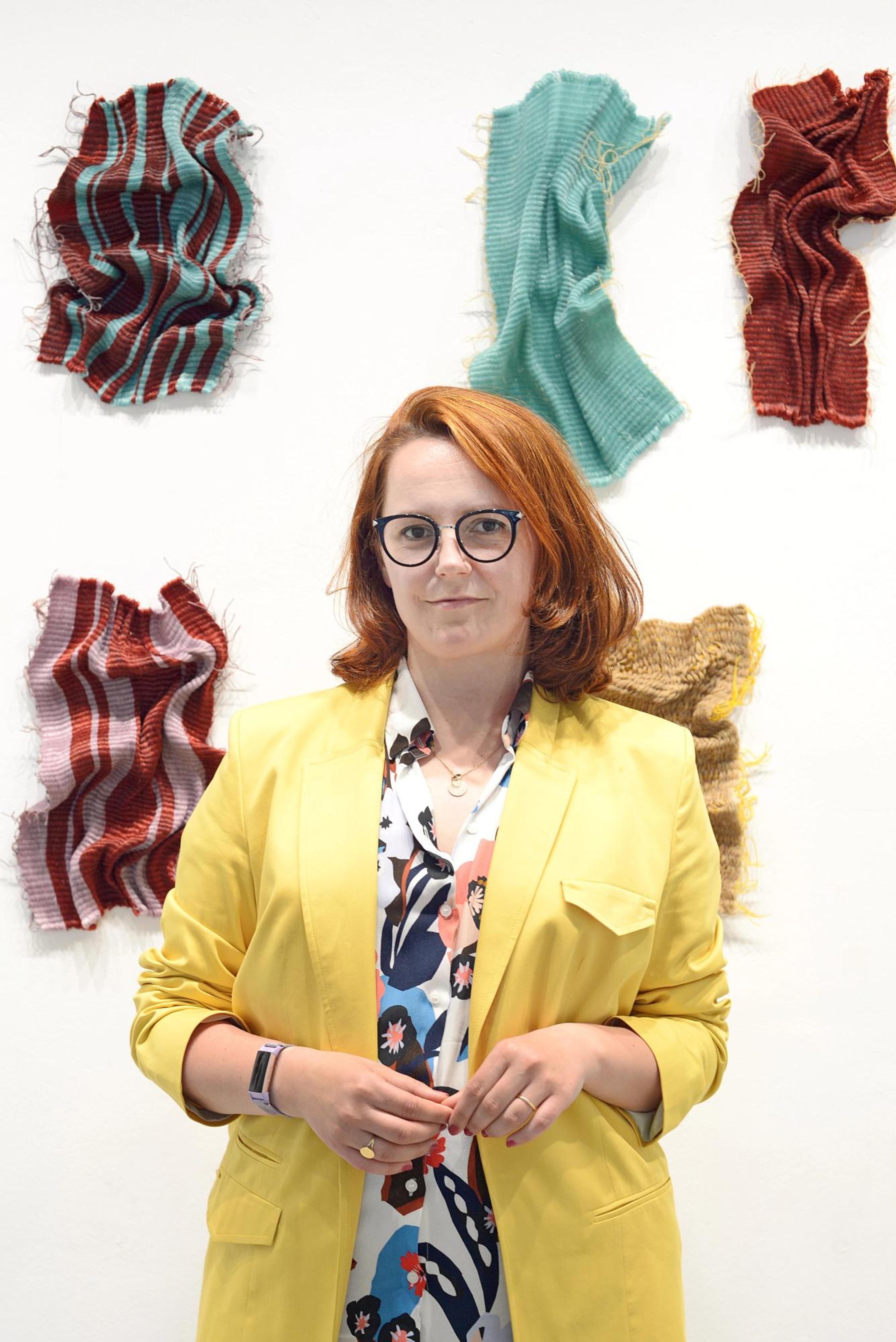 A woman in a yellow jacket stand in the frame. She has should length, red hair and wears a pair of black glasses. The background is a white wall with numerous textile artworks.