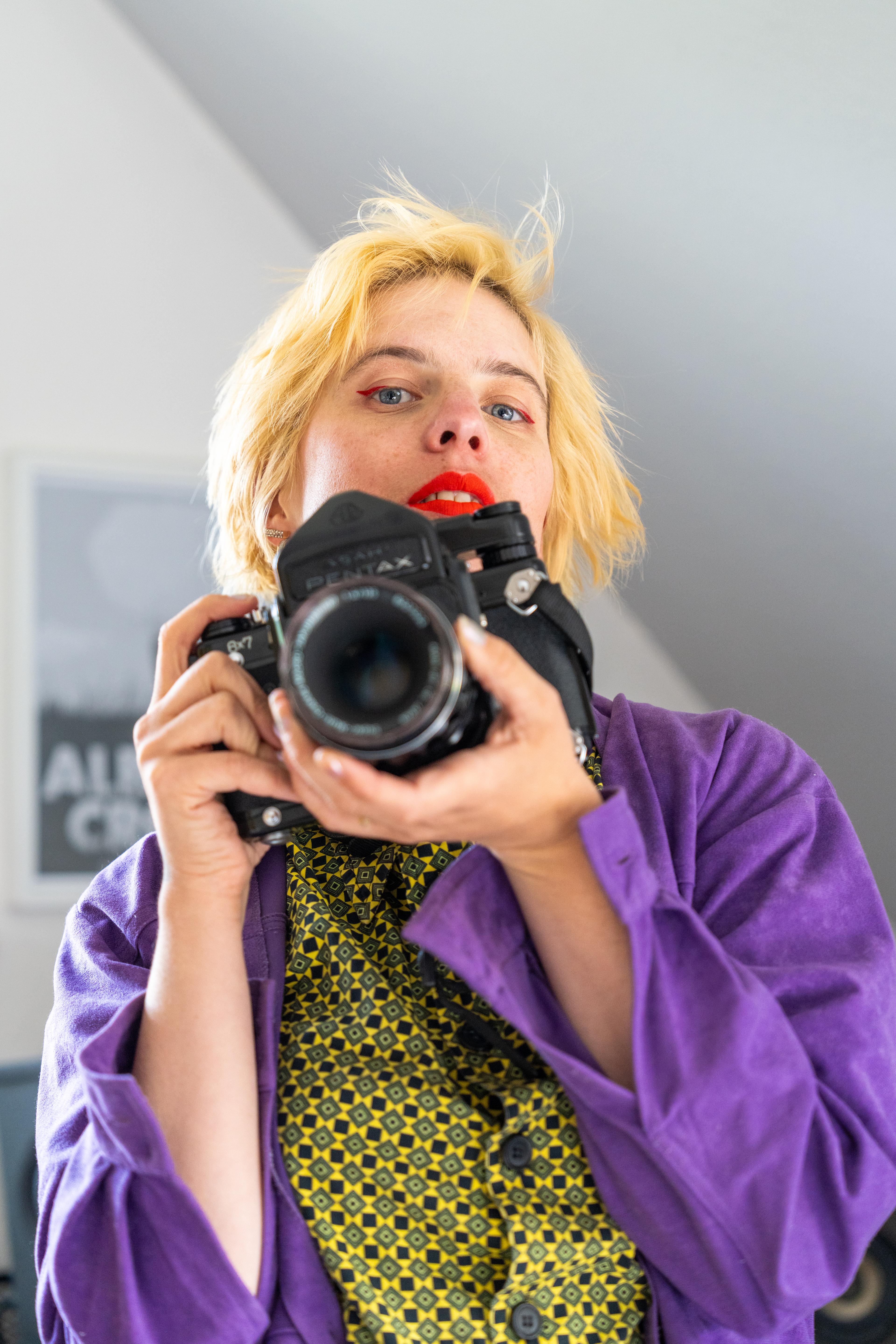 The artist looks to the viewer. She holds a large camera in her hand. She has short blonde hair and wears a purple jacket. Underneath is a green and yellow, checkered shirt.