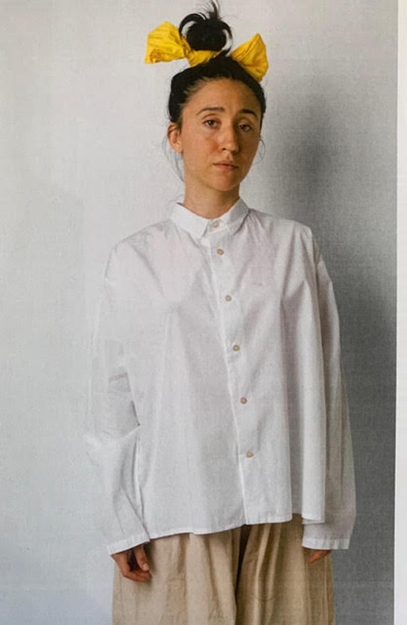 The artist Brigette Hoffman stands in front of a white wall. She is wearing a white long sleeve shirt, light brown trousers and has her hair in a bun. The hair is held together by a yellow band. 