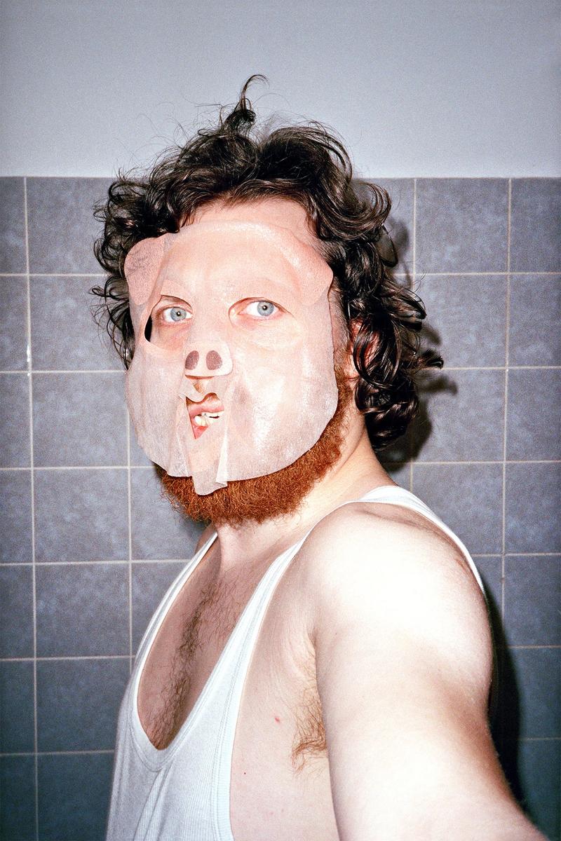 Portraitfoto Steve Braun, Mann im Unterhemd vor grauen Fließen mit Locken, einem roten Bart und einer Schwein-Gesichtsmaske