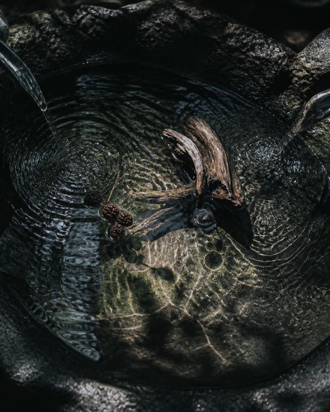 Two streams create overlapping ripples around wood and seed pods in a stone basin.
