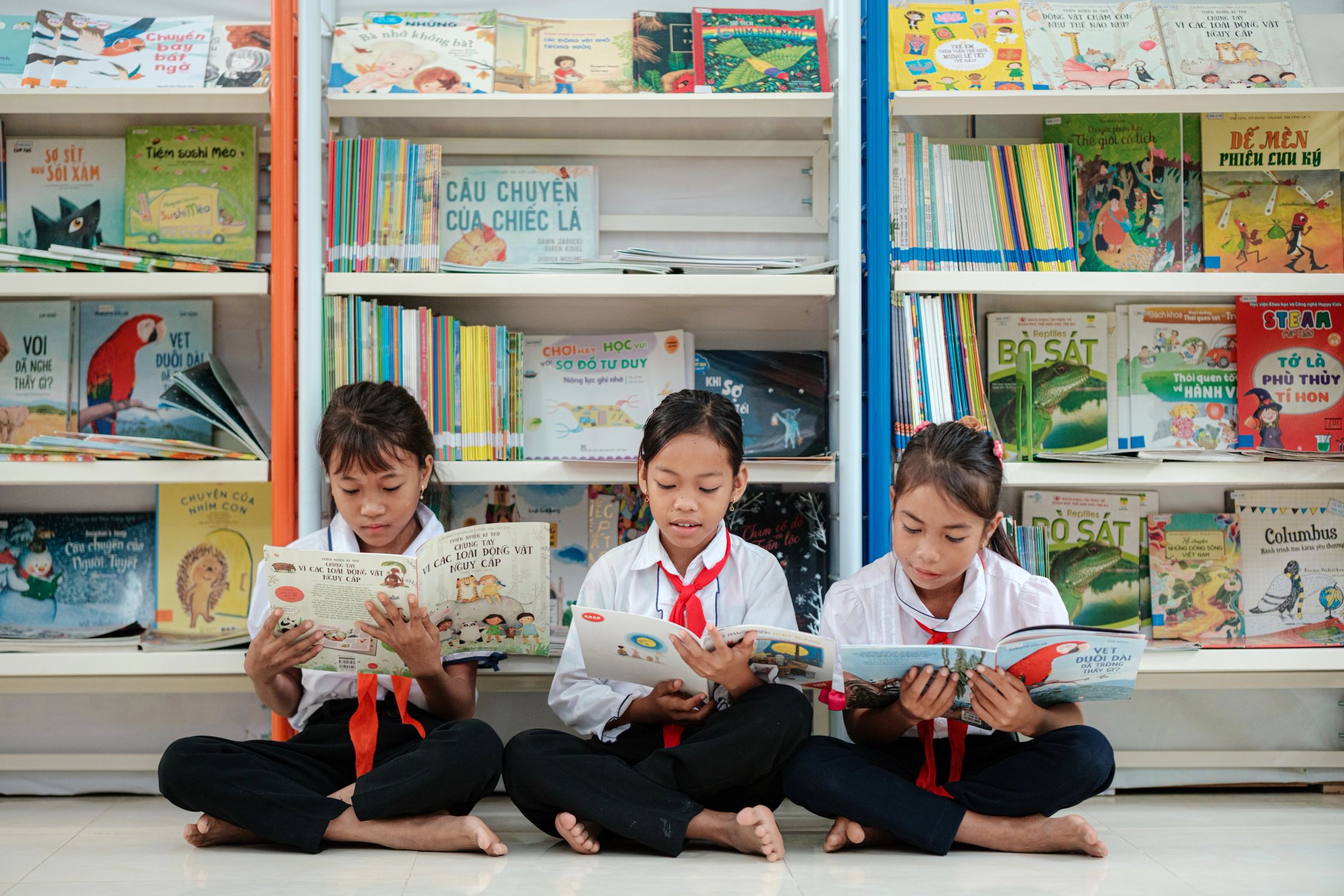 Three students sitting cross legged and reading books in front of a full bookshelf