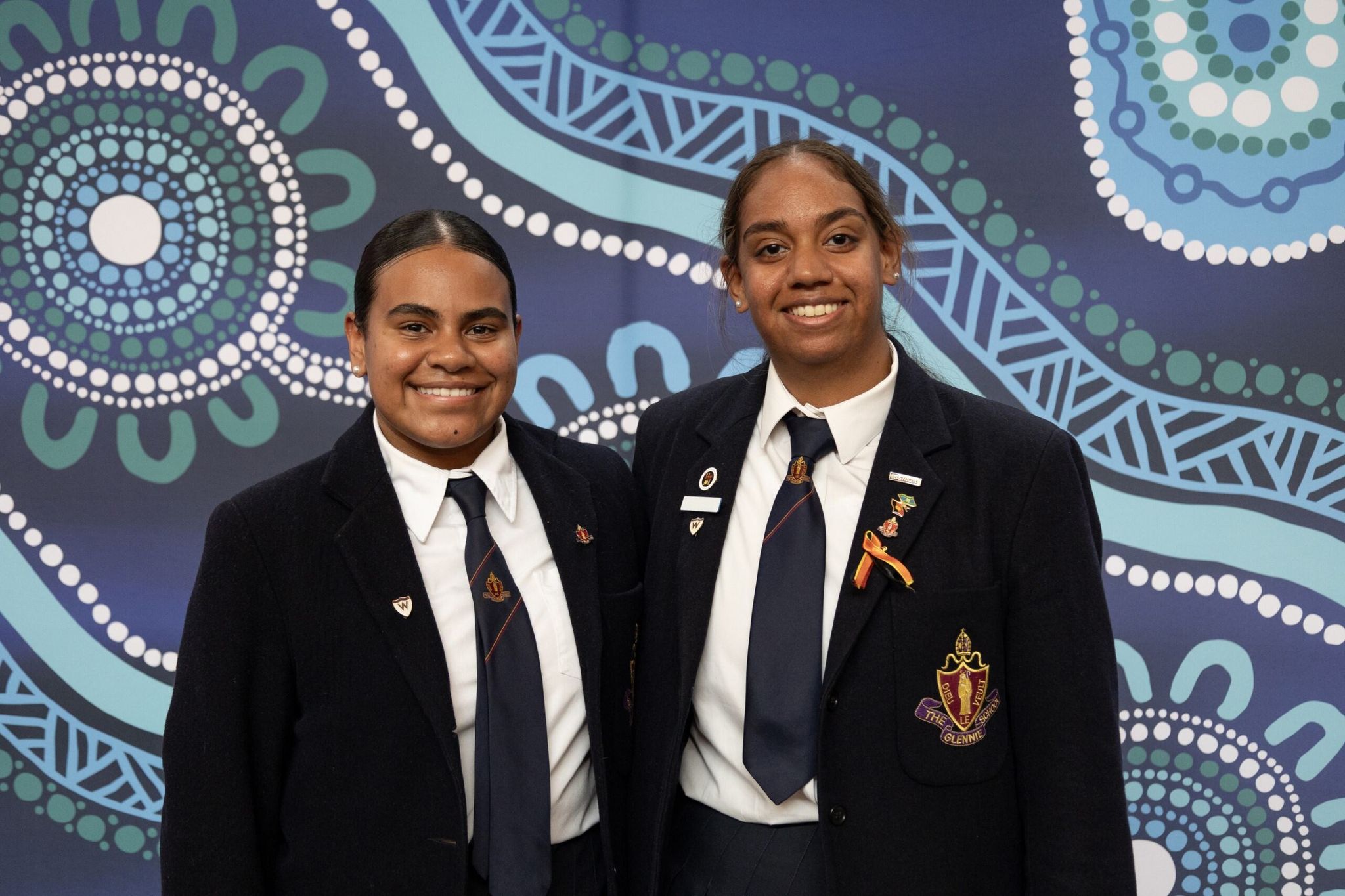 Two Yalari scholars in their school uniforms smiling against a blue background with aboriginal art patterns