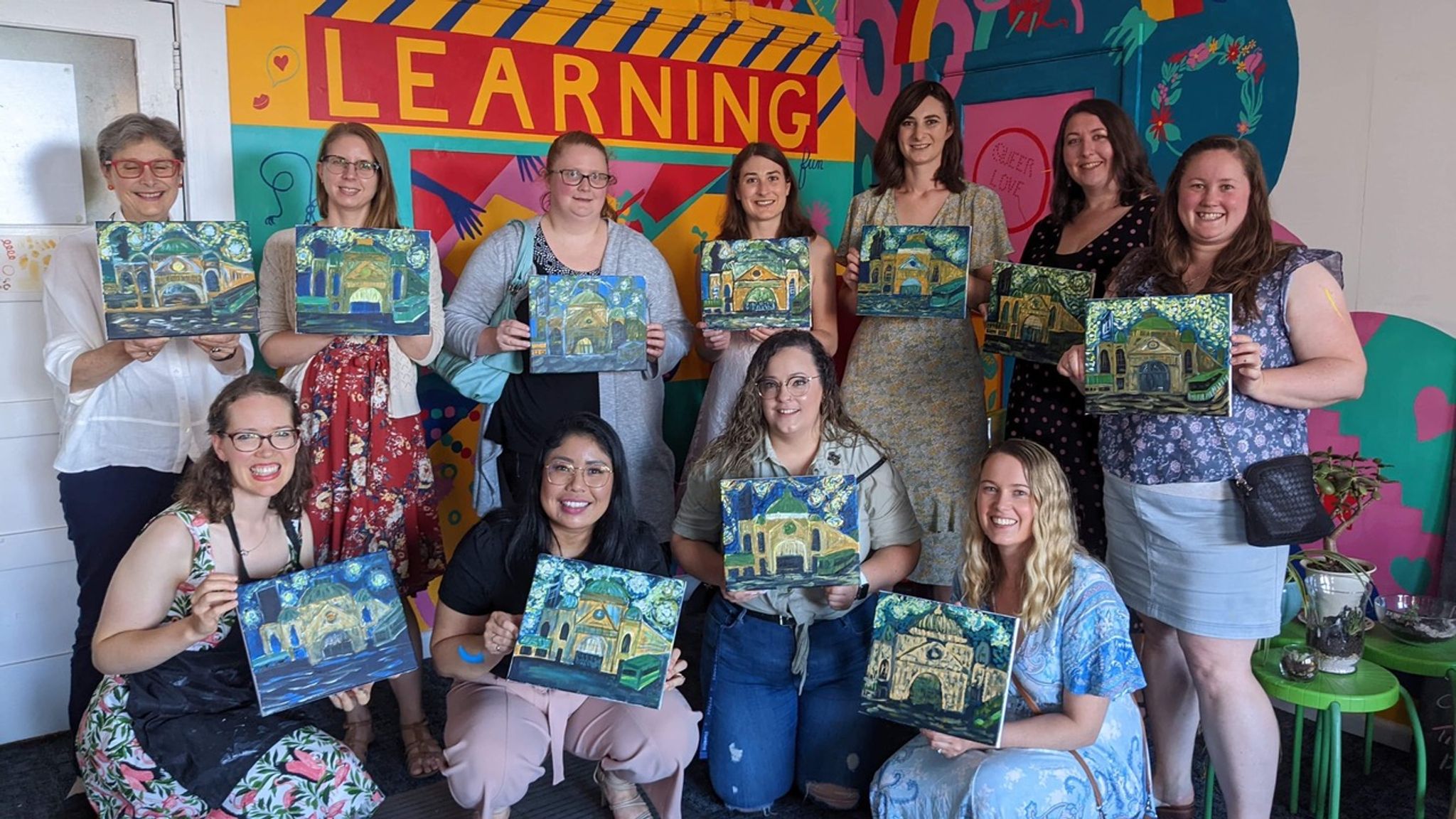 Students holding up paintings with a colourful mural in the background with words 'learning'