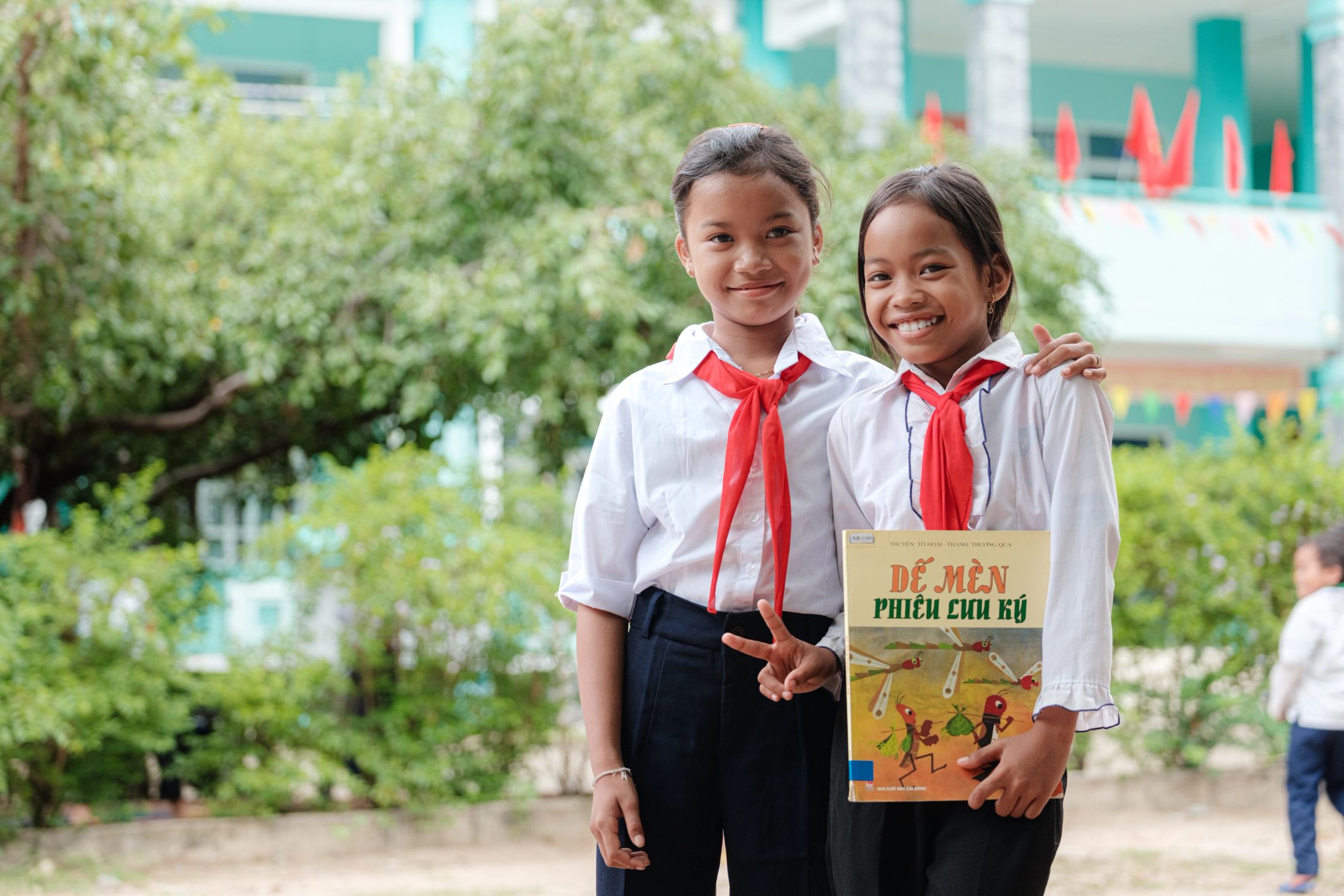 Two students smiling and holding a book in front of a school in Vietnam