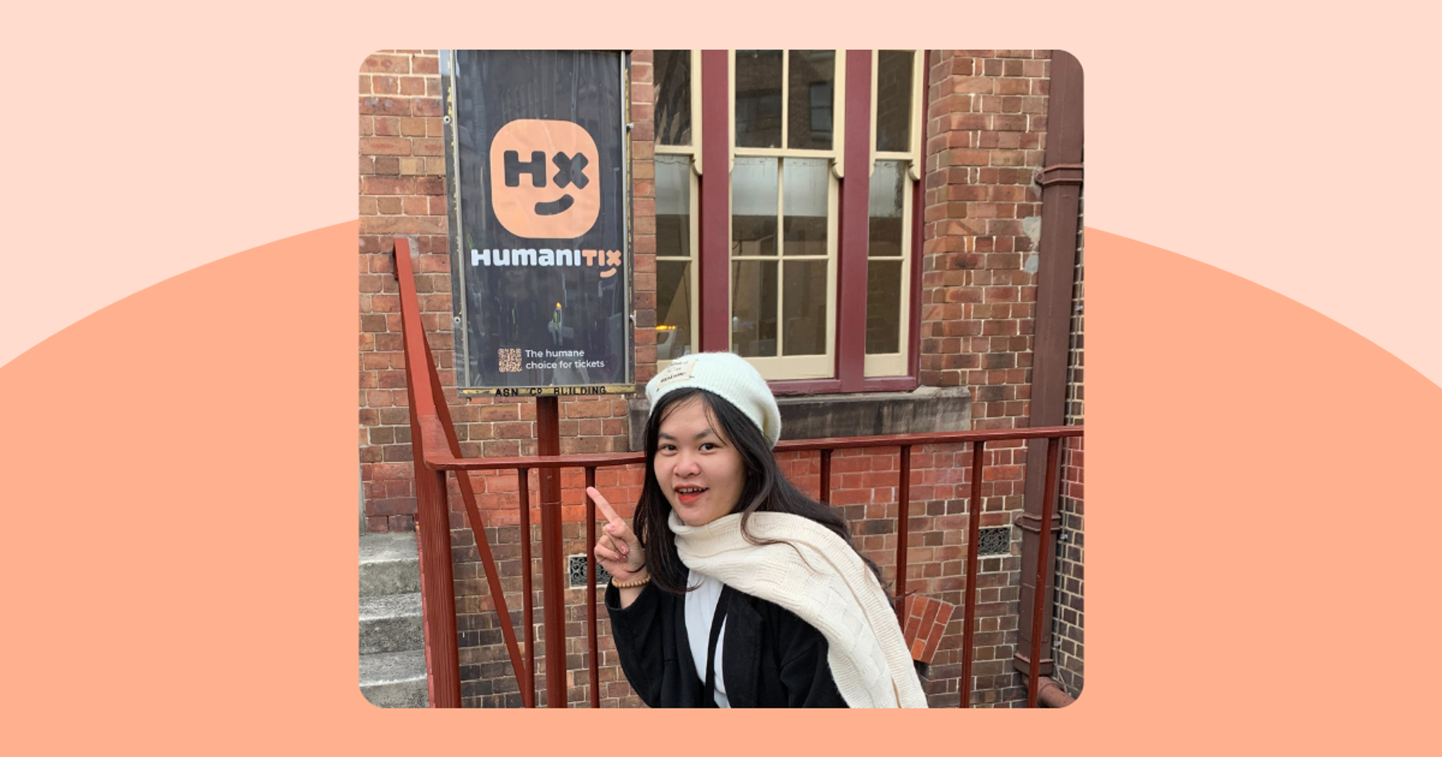 A girl in a black coat and beige hat an scarf points at a sign showing the Humanitix logo