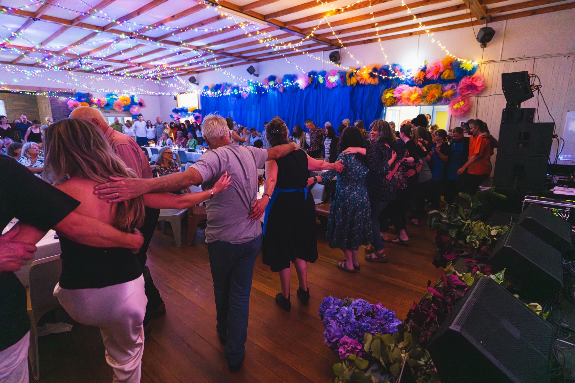 Festival-goers standing in a line holding each others shoulders and waist in a room lit up by fairy lights