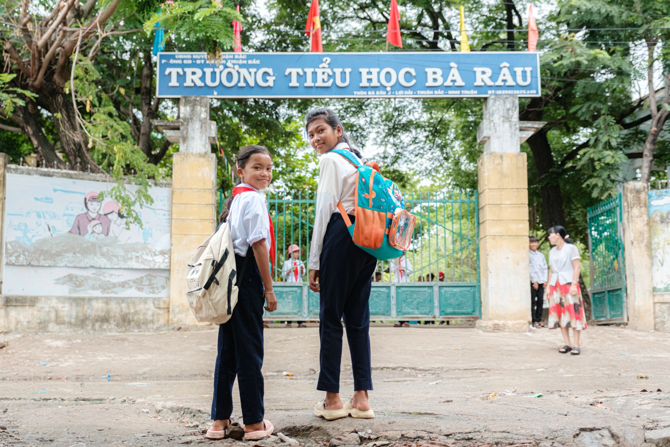 Two students stand with backpacks in front of the Truong Tieu Hoc Ba Rau school sign