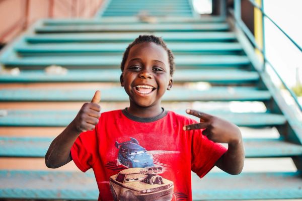 A smiling young African boy doing a thumbs up and a peace sign