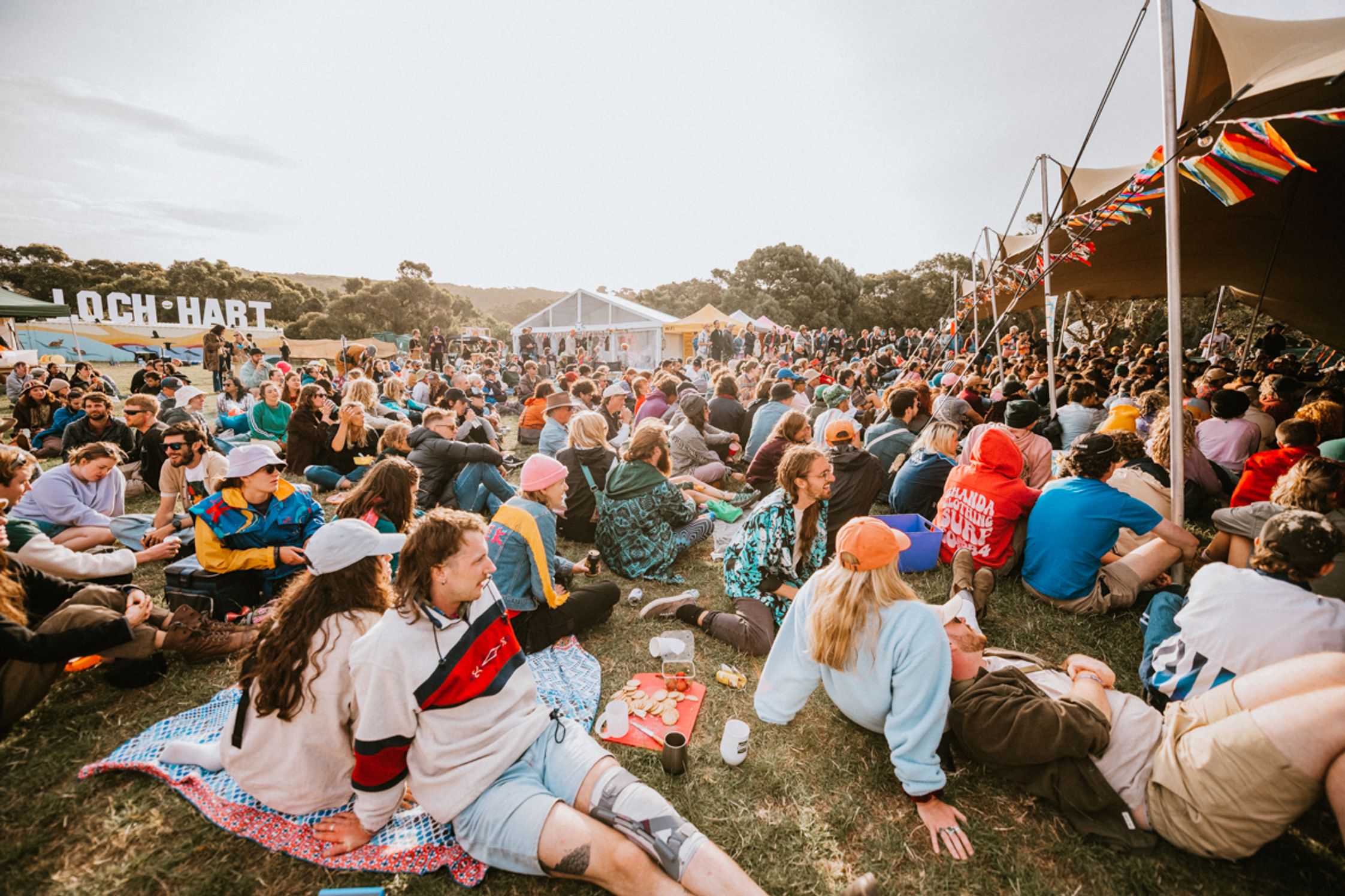 Hundreds of festival attendees sitting in the sun in the grass at Loch Hart festival