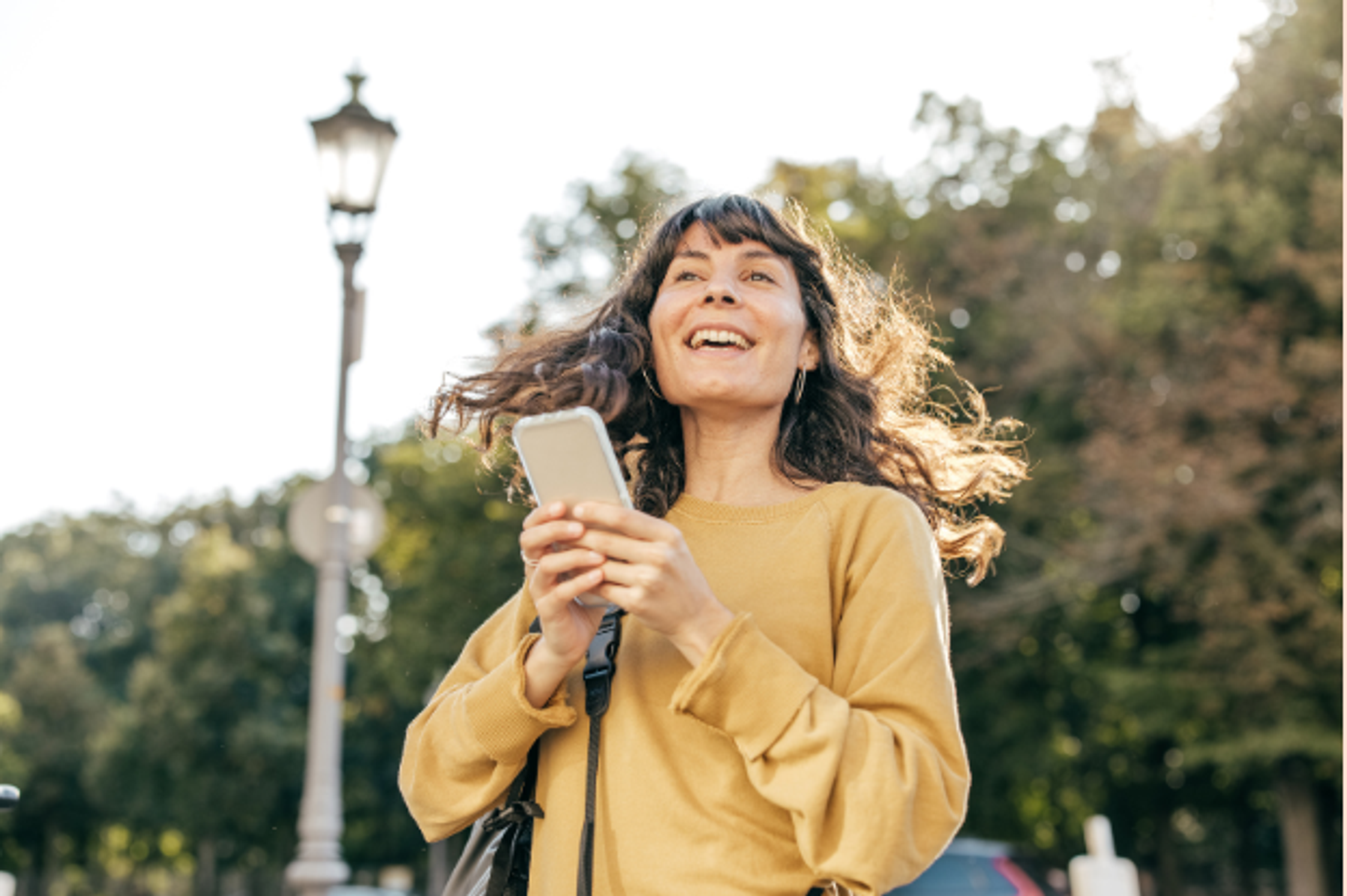 A woman in yellow sweater holds her phone and is smiling into the distance