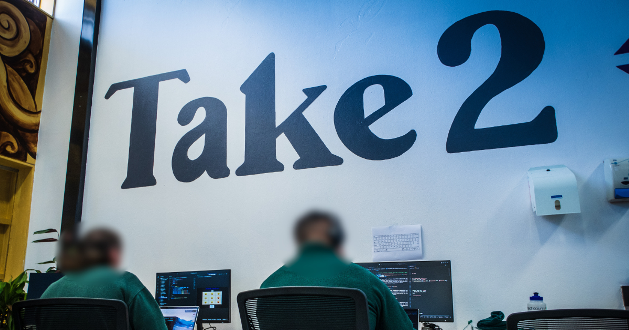 The Take2 logo on a wall with two inmates coding in the foreground