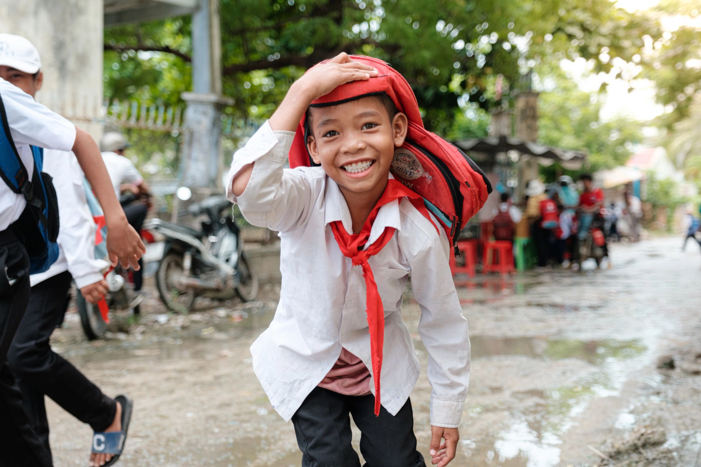 A smiling boy in school uniform holding a bag over his head in Vietnam to avoid the rain