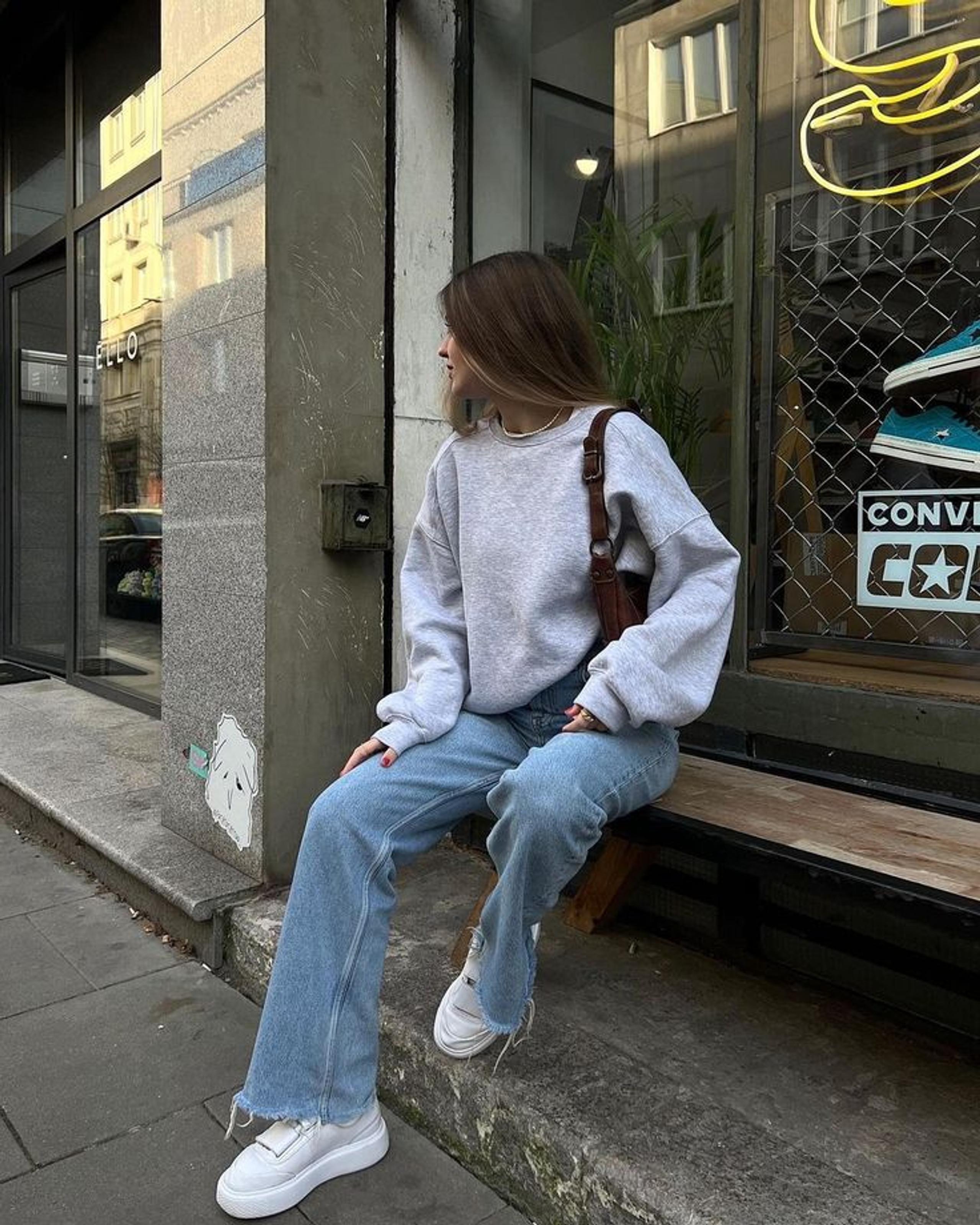 Woman in an oversized grey crewneck jumper, straight-leg blue jeans and brown loafers walking down a London street