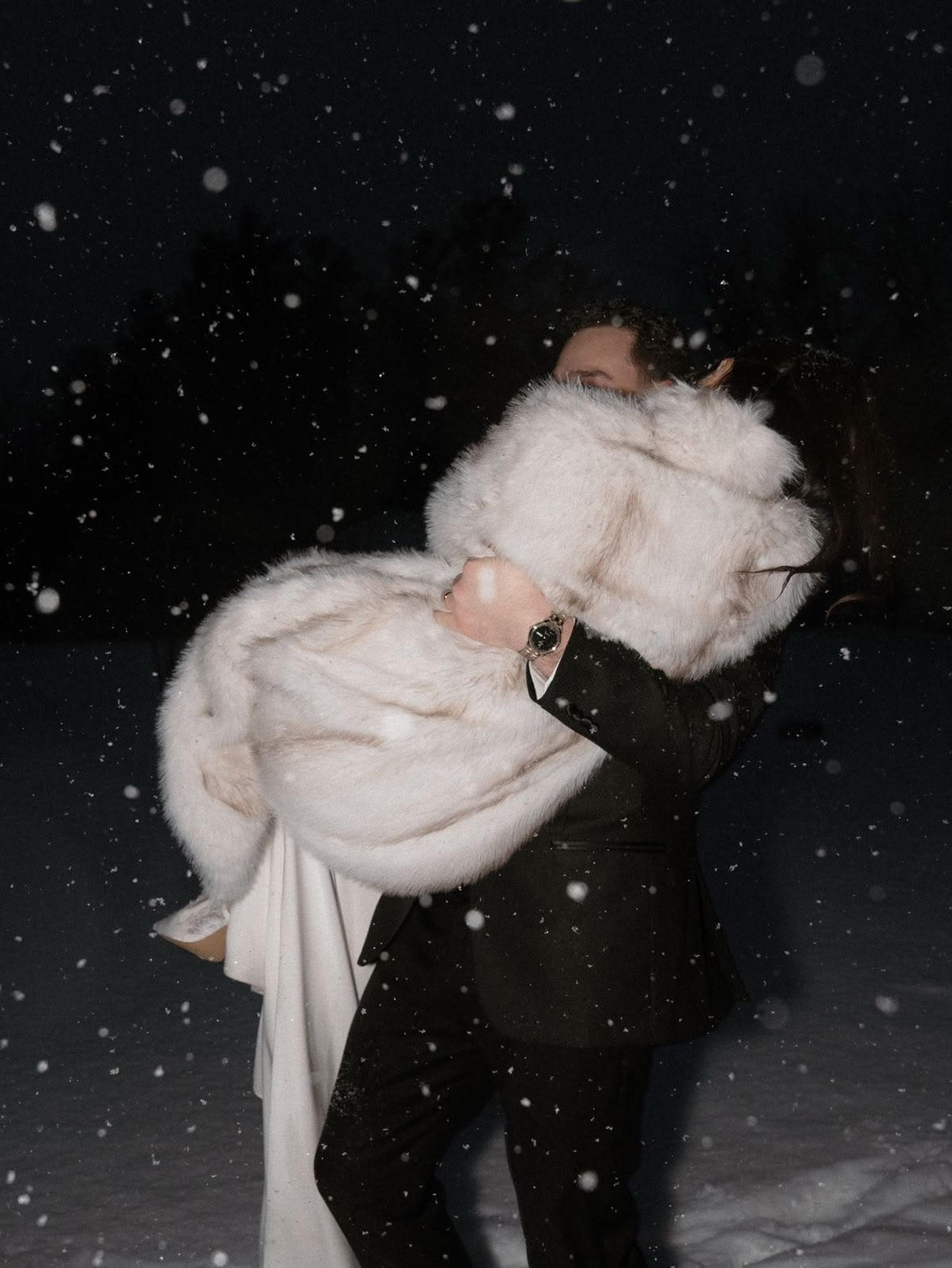 Bride in a white faux fur coat being carried through falling snow at night