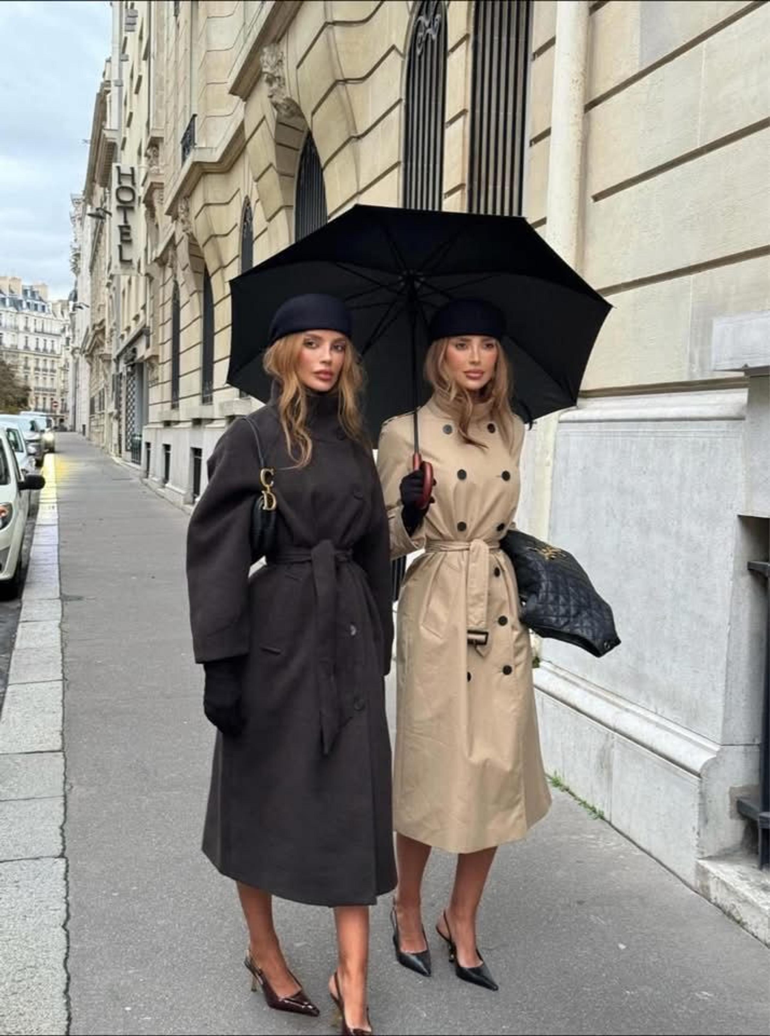 Two women under a black umbrella in a chocolate belted coat and a classic beige trench on a Paris street