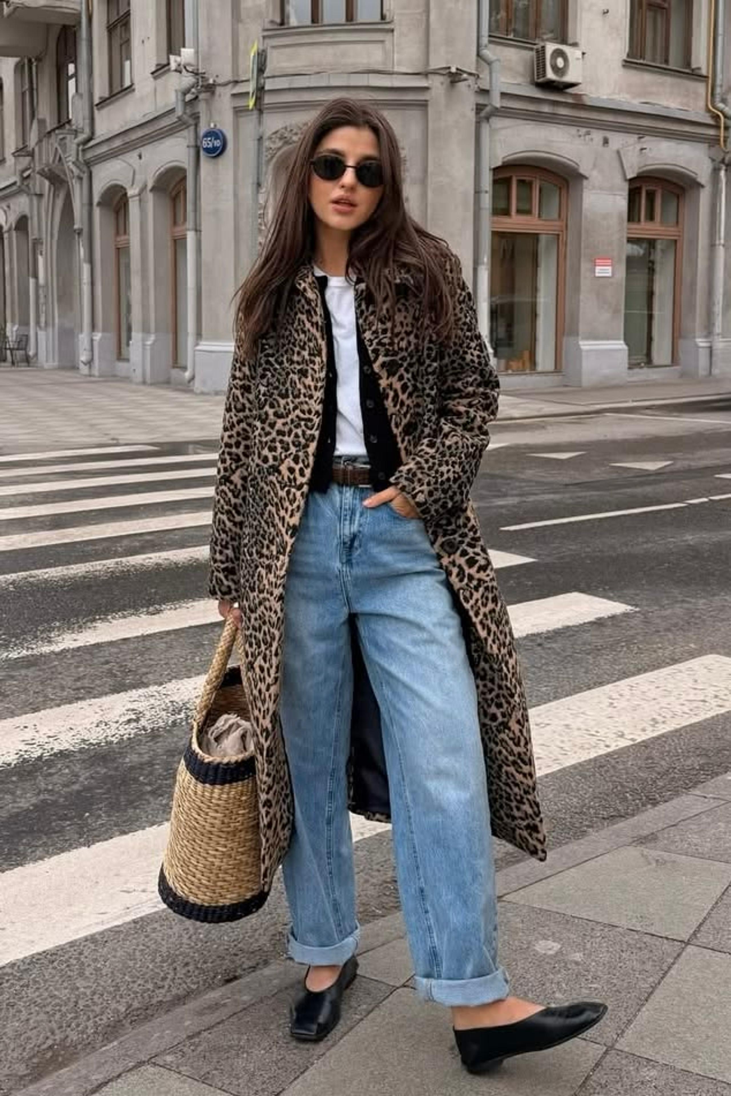Woman in a long leopard coat, white tee, black cardigan and turn-up jeans with a straw bag on a city crossing