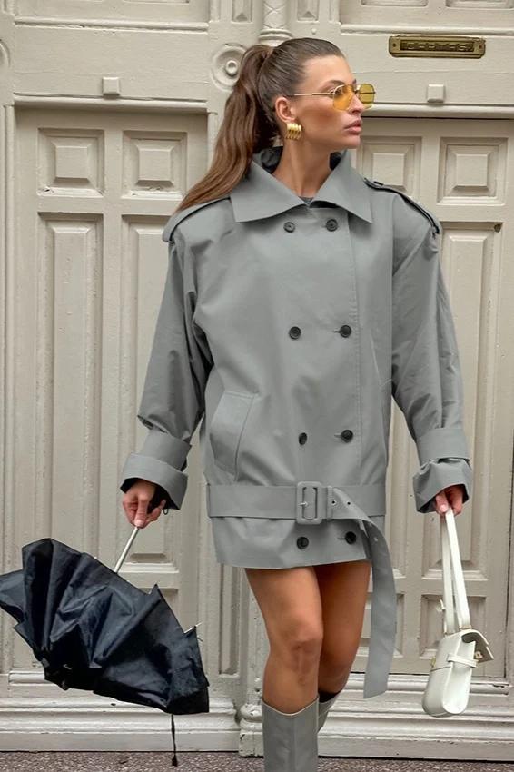 Woman in a short grey belted trench, gold earrings and white bag carrying a black umbrella