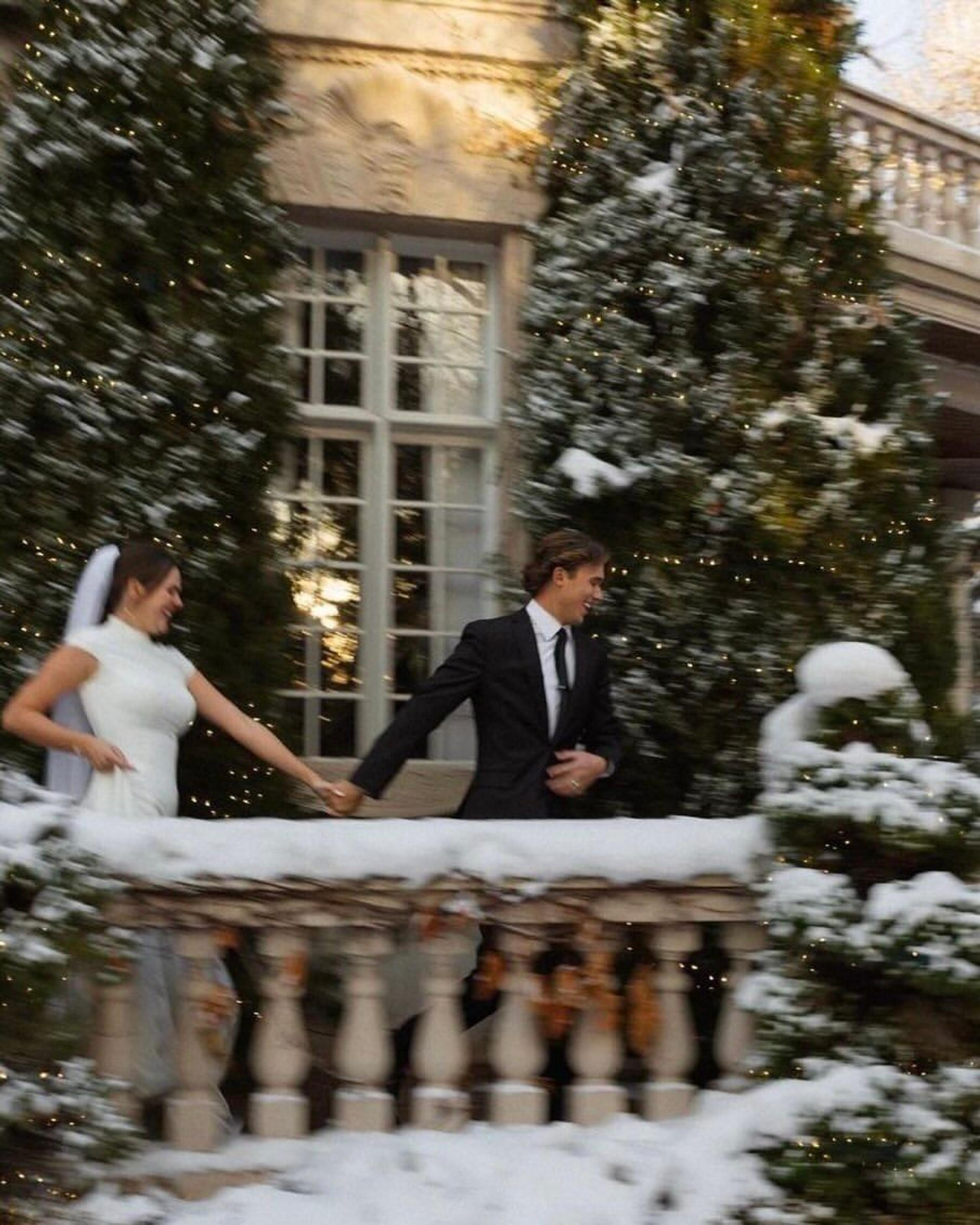Bride in a white gown and groom in a black suit on a snowy balcony with fir trees
