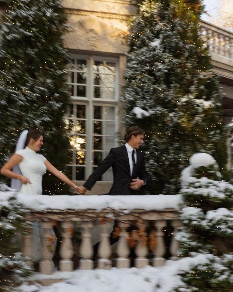 Bride in a white gown and groom in a black suit on a snowy balcony with fir trees