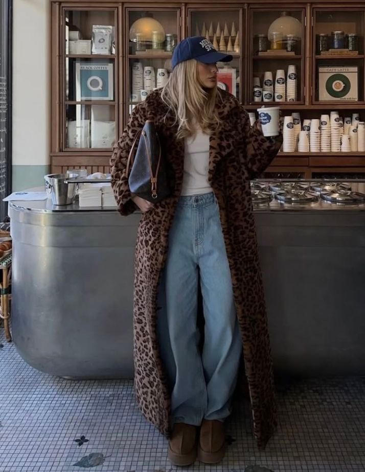 Woman in a long leopard-print coat, baggy jeans and a navy cap standing in a deli