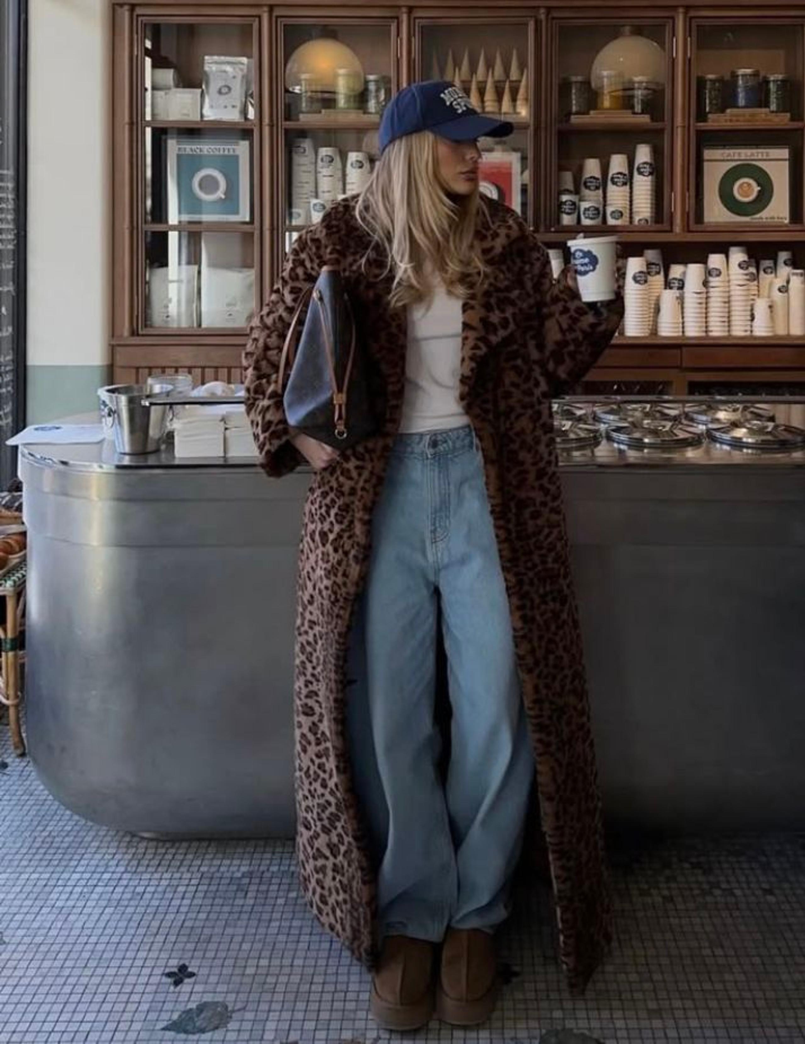 Woman in a long leopard-print coat, baggy jeans and a navy cap standing in a deli