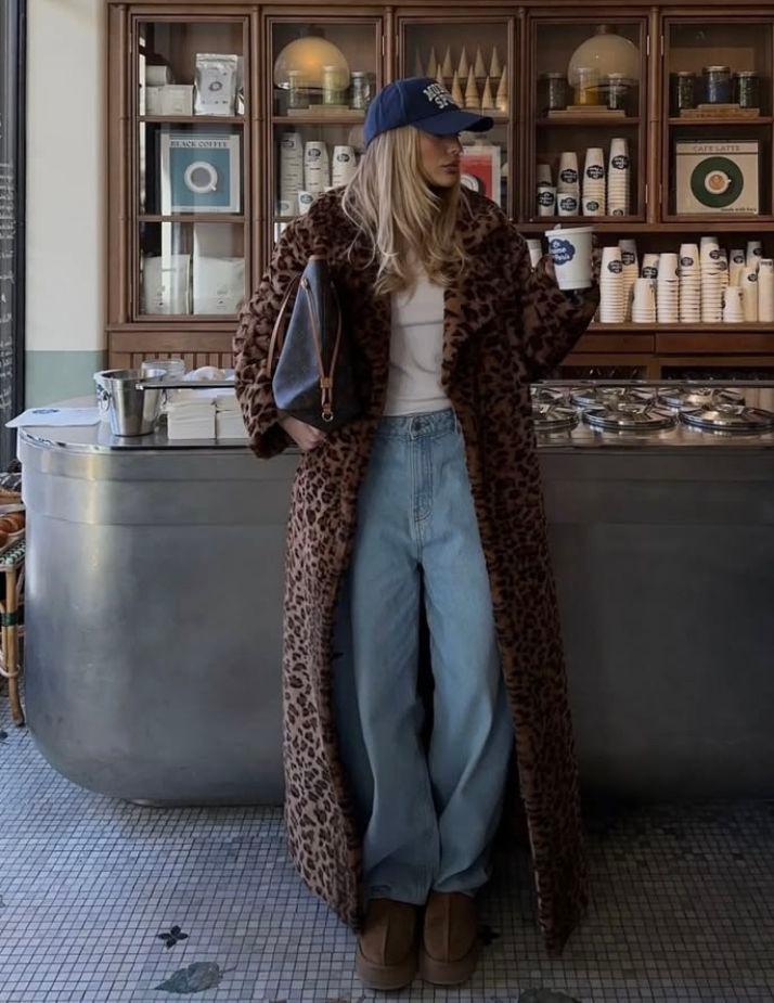 Woman in a long leopard-print coat, baggy jeans and a navy cap standing in a deli