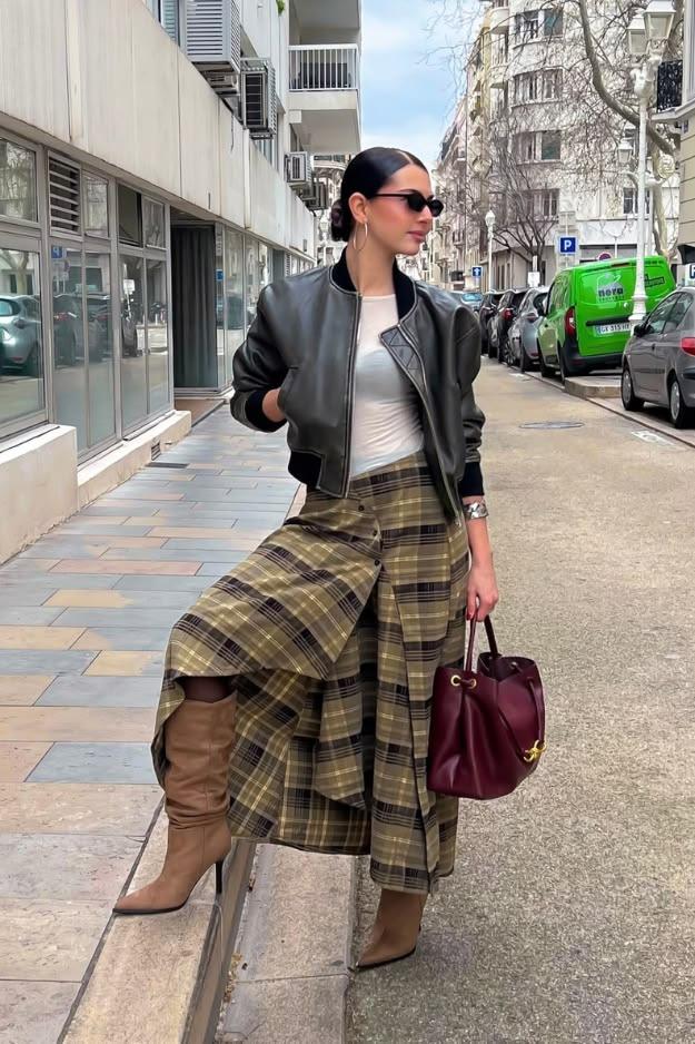 Woman in a long olive tartan skirt, black leather bomber, tan boots and burgundy bag on a city pavement