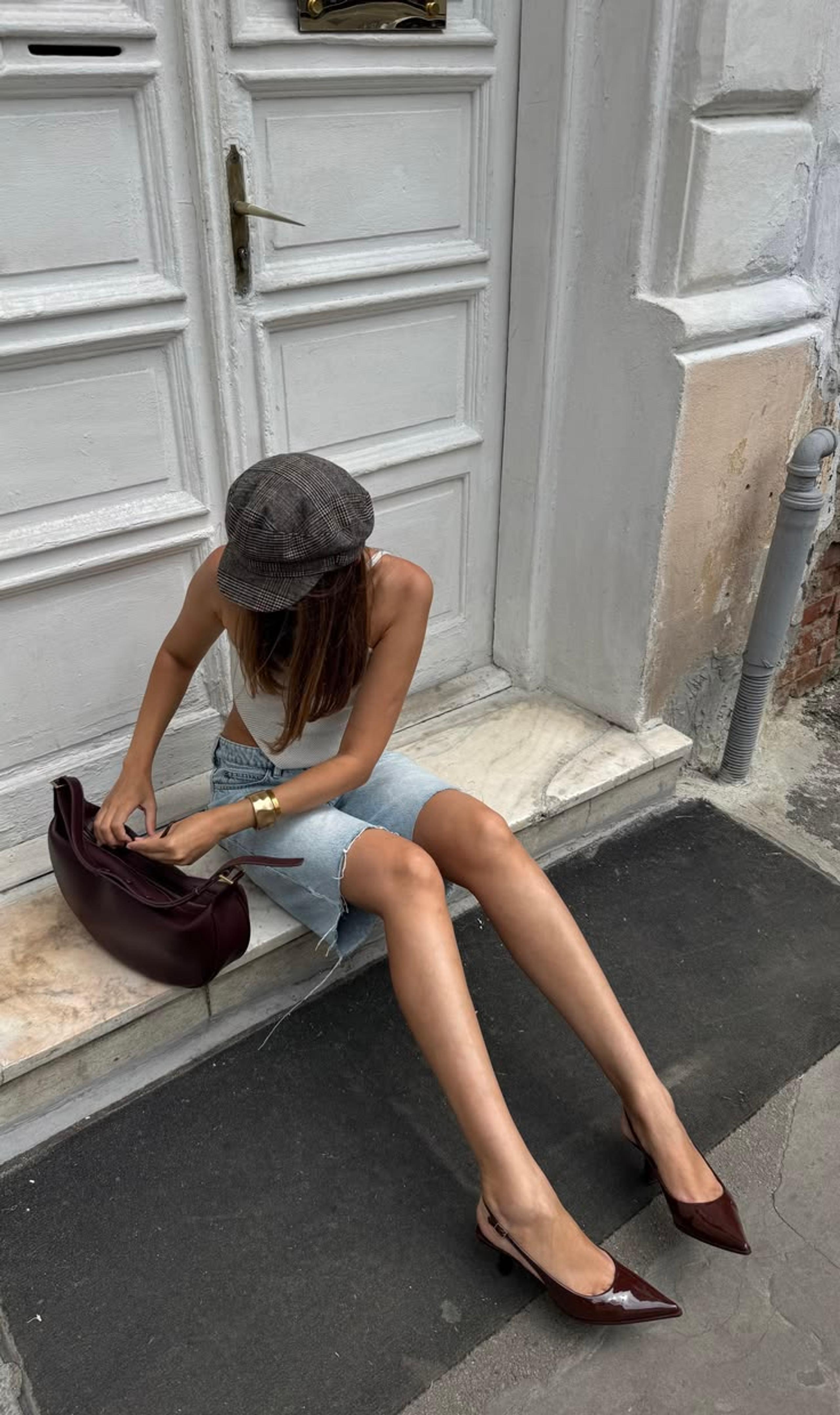 Woman in a white vest, denim shorts and brown pointed heels with a burgundy slouch bag sitting on a step